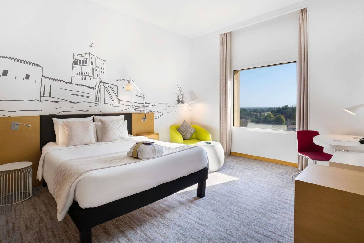 A hotel room with a bed, a desk, a chair, a lamp, a window with a view, and a wall mural.