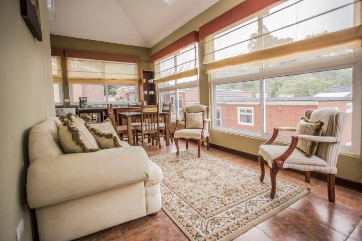 A spacious living room with a beige sofa, two armchairs, a patterned rug, and large windows offering a view of the neighborhood.