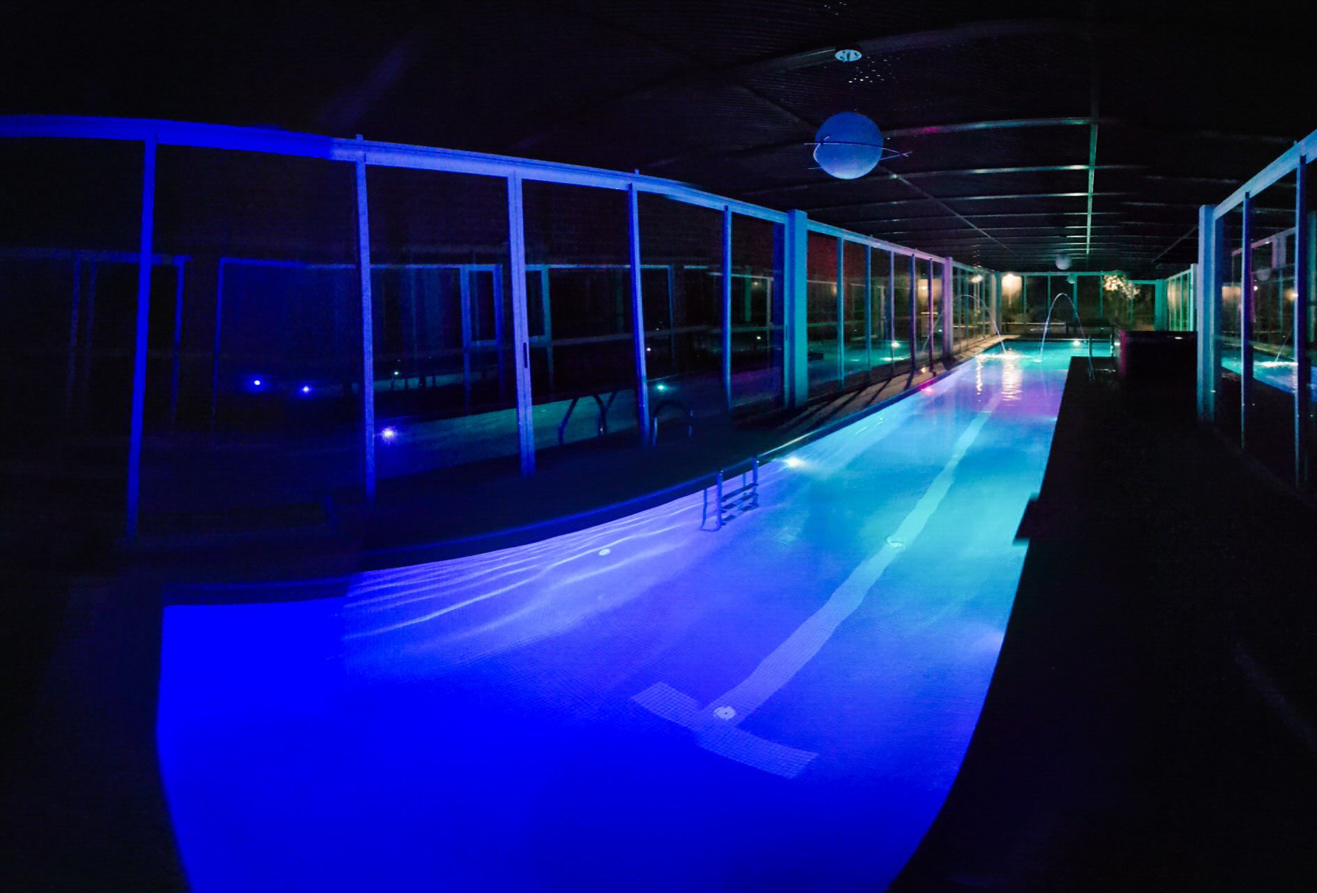 A swimming pool with a curved design, surrounded by glass walls and illuminated by blue lighting.