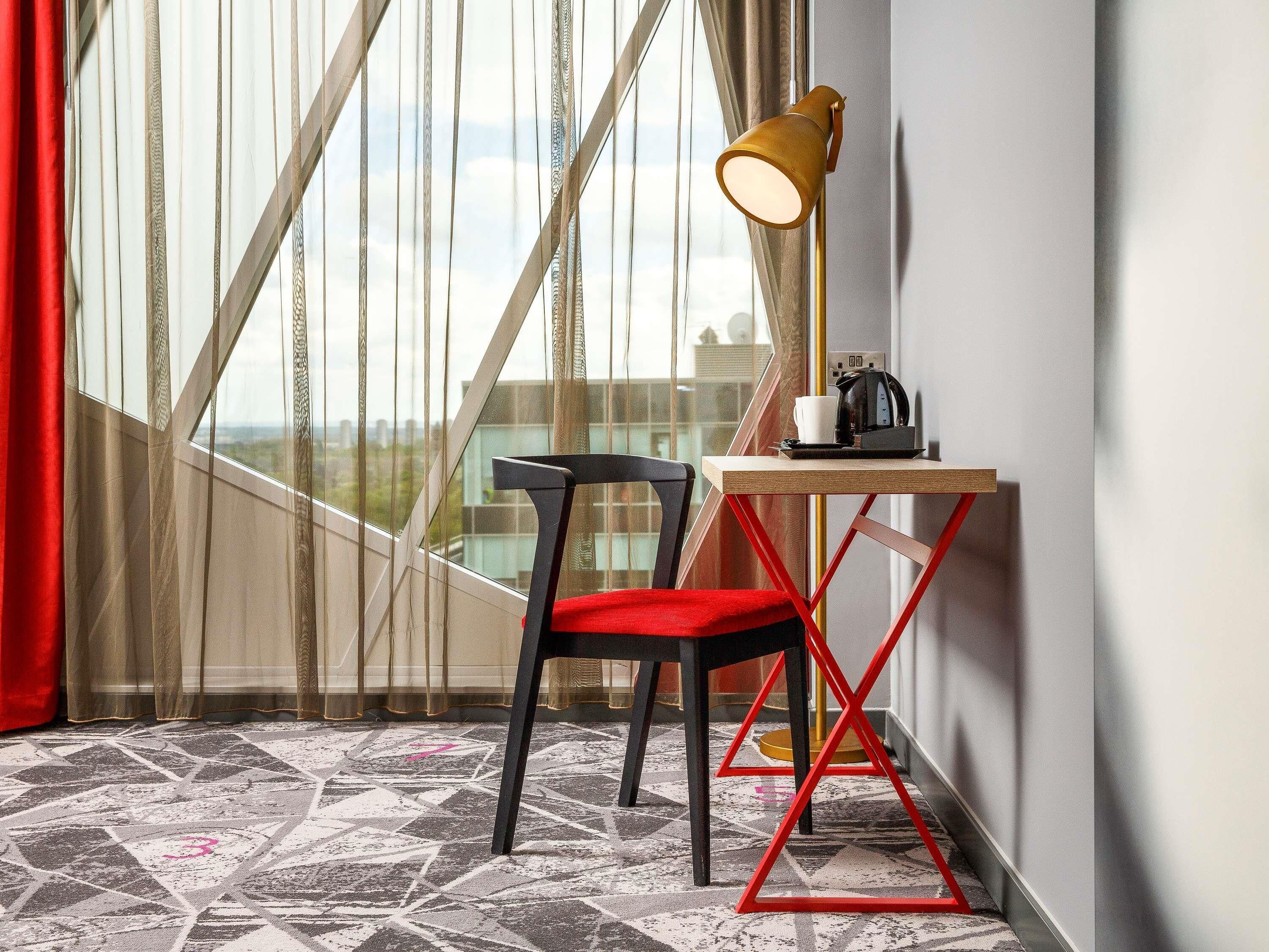 A room with a view through sheer curtains, a desk with a coffee maker, and a chair with a red cushion.