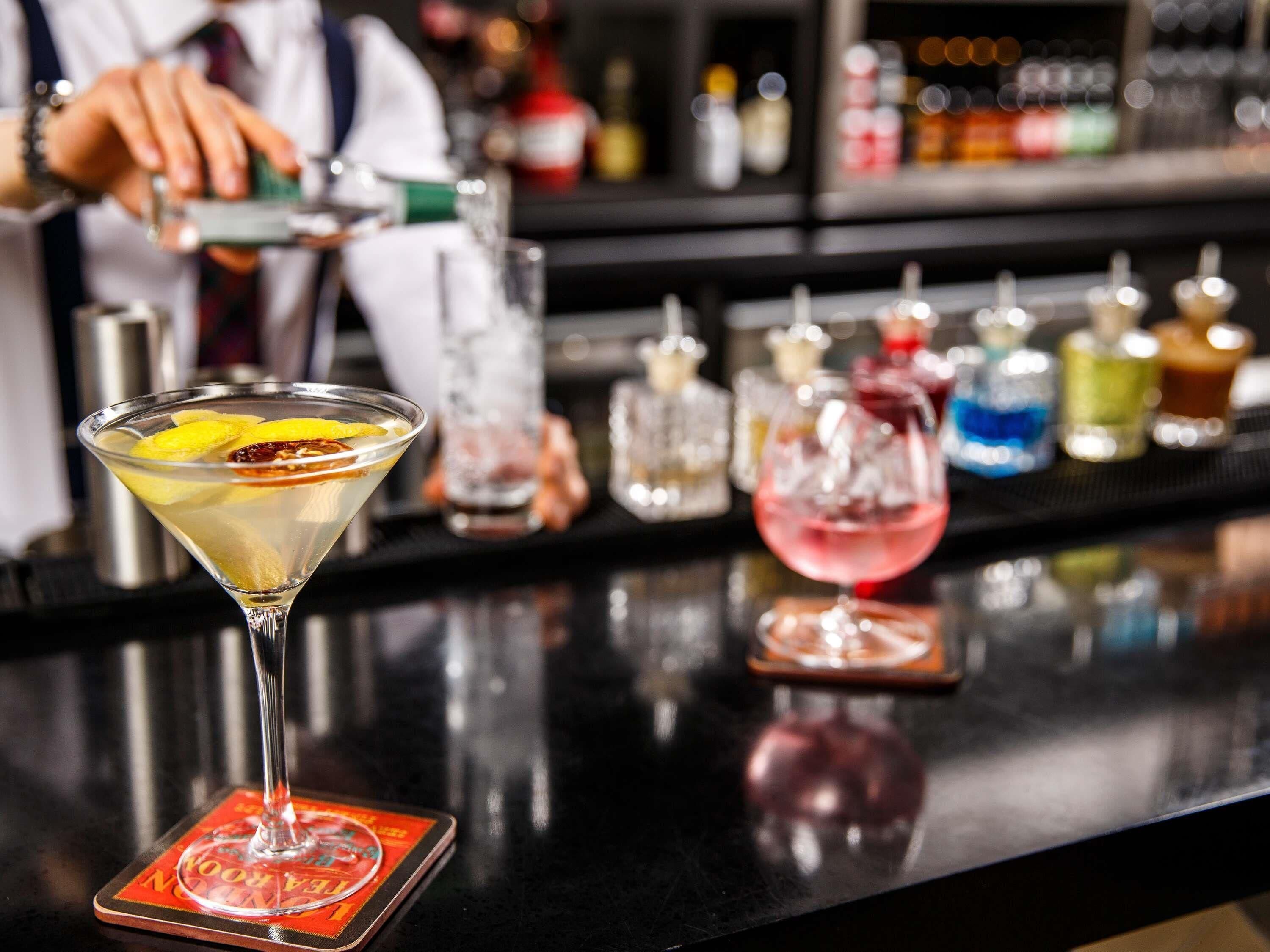 Una copa de martini con una rodaja de limón sobre un posavasos, un bar con varias botellas y copas al fondo.