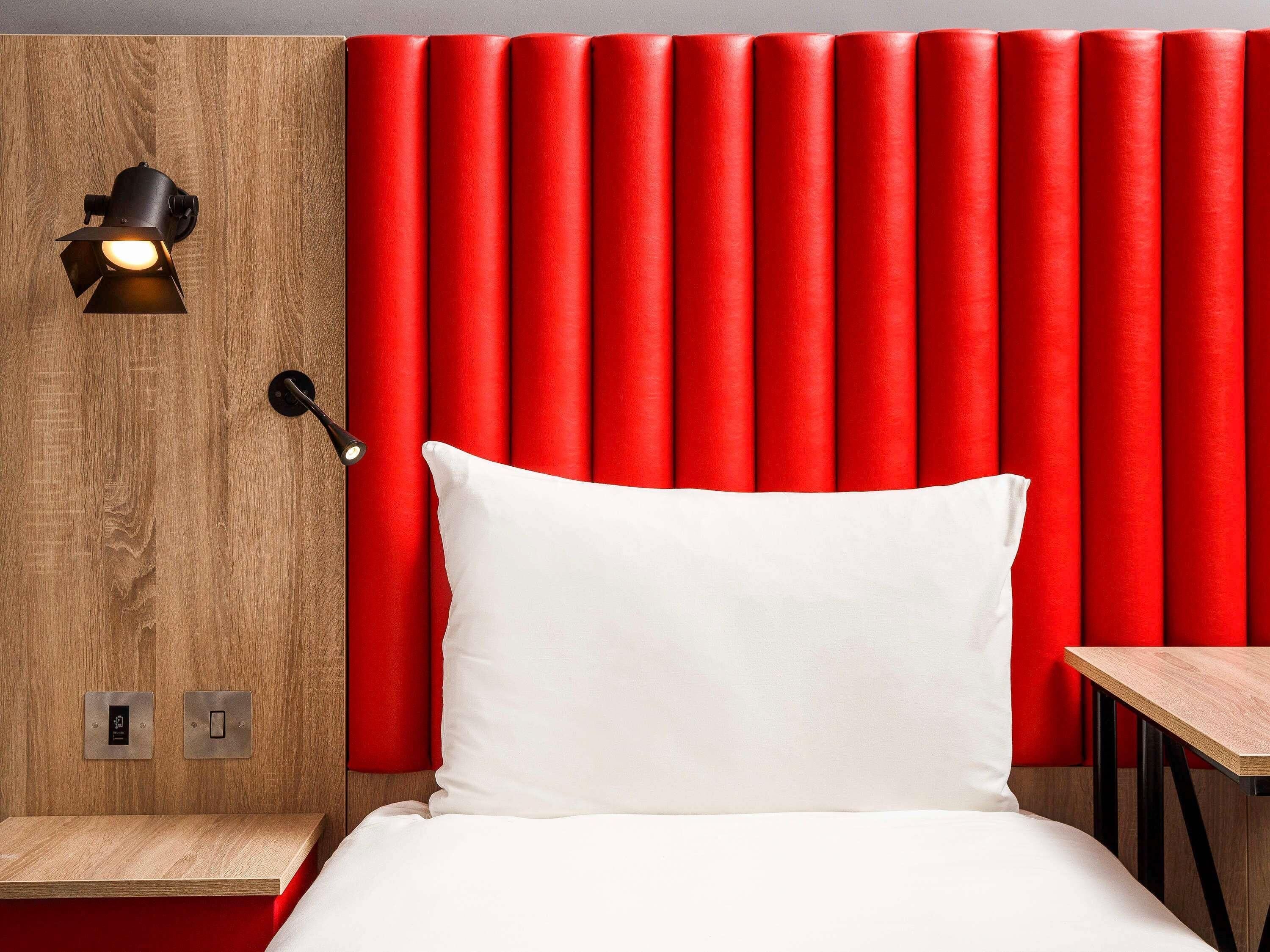 Une chambre d’hôtel avec une tête de lit en bois, un oreiller blanc, une table de chevet et un mur d’accent rouge.