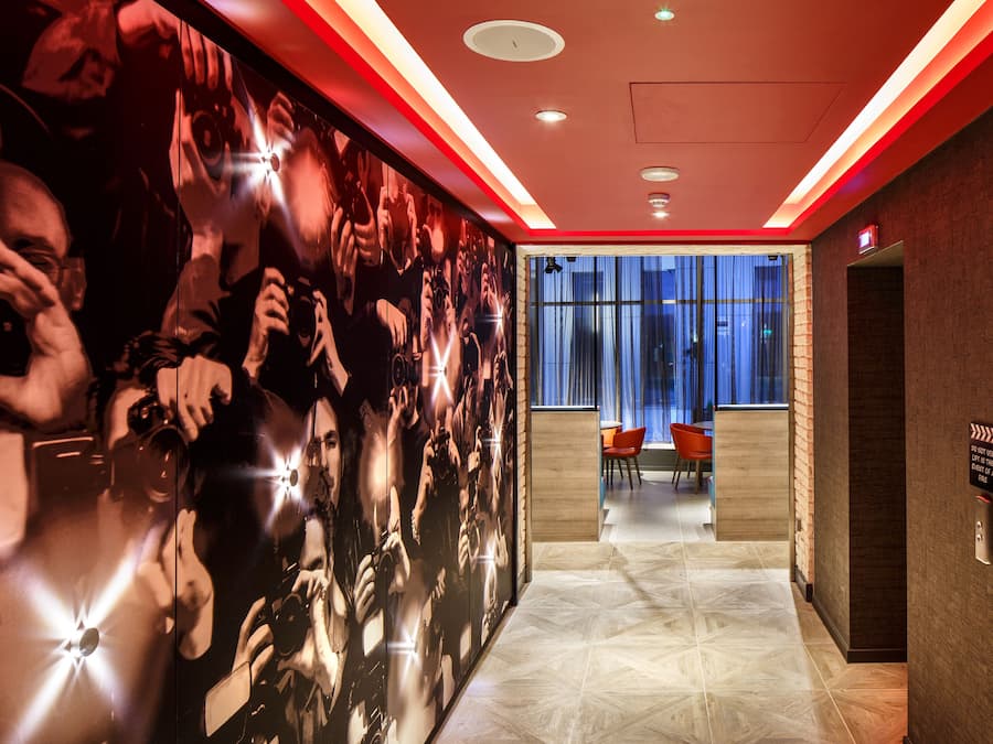 A hallway with a mirrored wall reflecting people taking photos, a sign indicating no smoking, and a view into a room with a blue curtain.