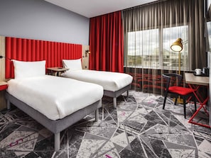 Une chambre dâhĂŽtel avec deux lits, un bureau, une chaise, une lampe et une vue sur la ville grĂące Ă de grandes fenĂȘtres.