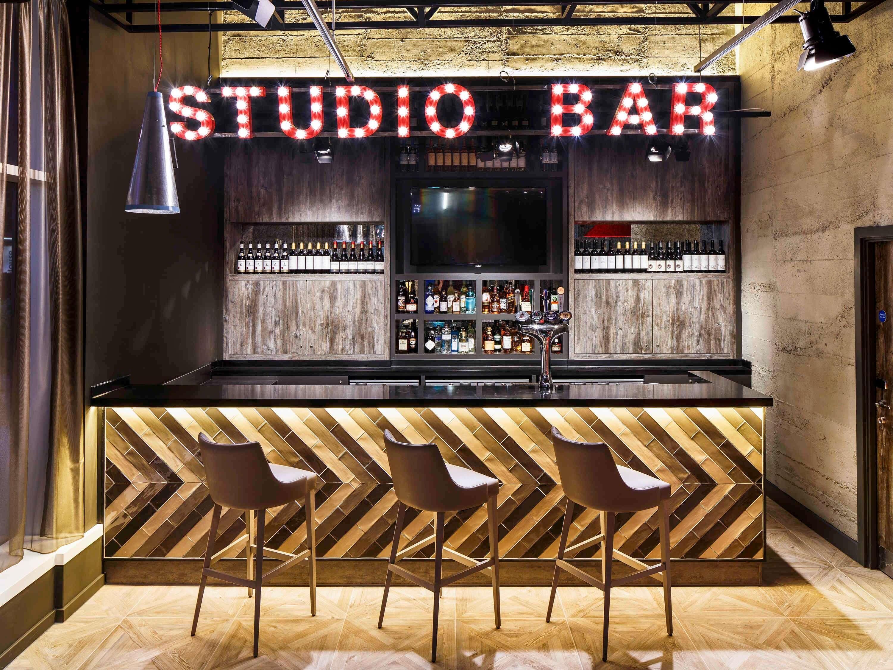 A bar area with a wooden herringbone floor, three bar stools, and a variety of bottles and glasses on shelves.