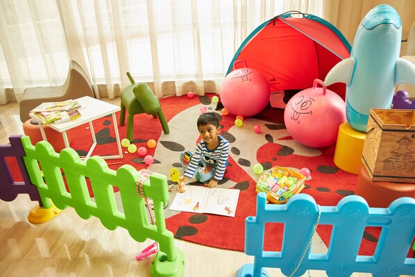 Un niño sentado sobre una alfombra roja con una carpa, pelotas de colores y una mesa de juguetes en una sala de juegos.