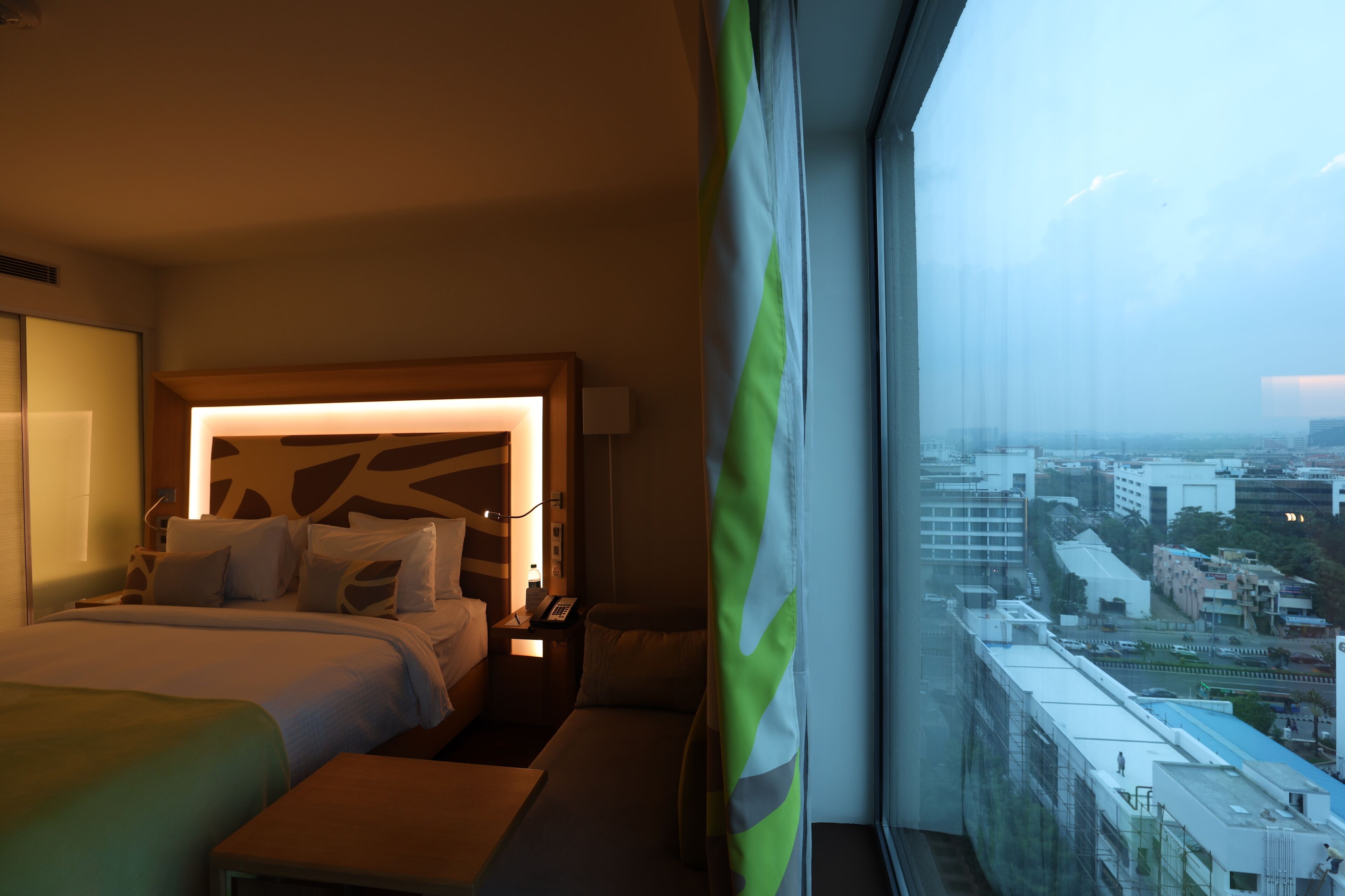 Habitación de hotel con una cama grande, mesitas de noche, un escritorio y vistas a la ciudad a través de un amplio ventanal.