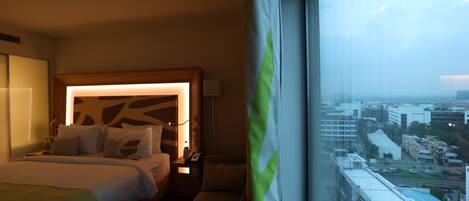 Habitación de hotel con una cama grande, mesitas de noche, un escritorio y vistas a la ciudad a través de un amplio ventanal.