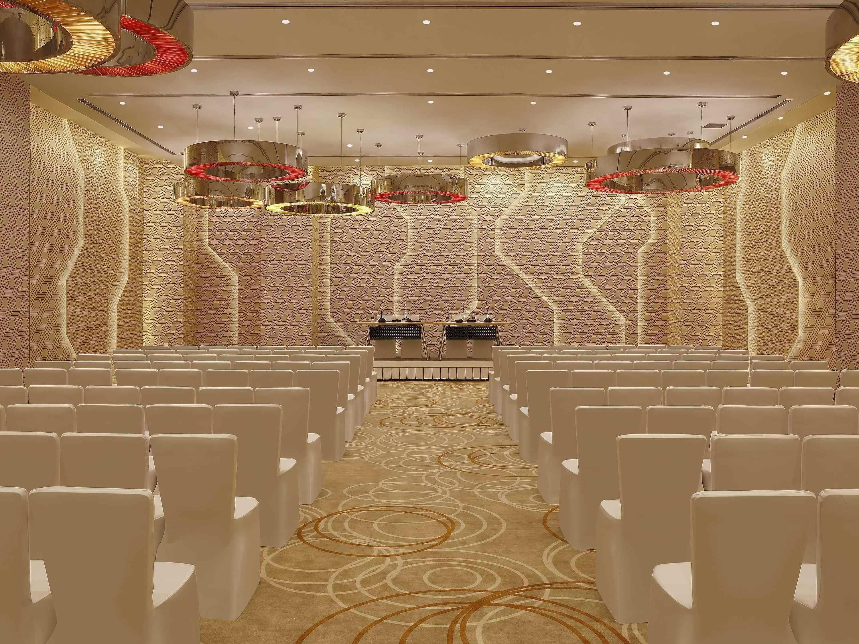 A conference room with rows of white chairs, a stage with a podium, and modern chandeliers.