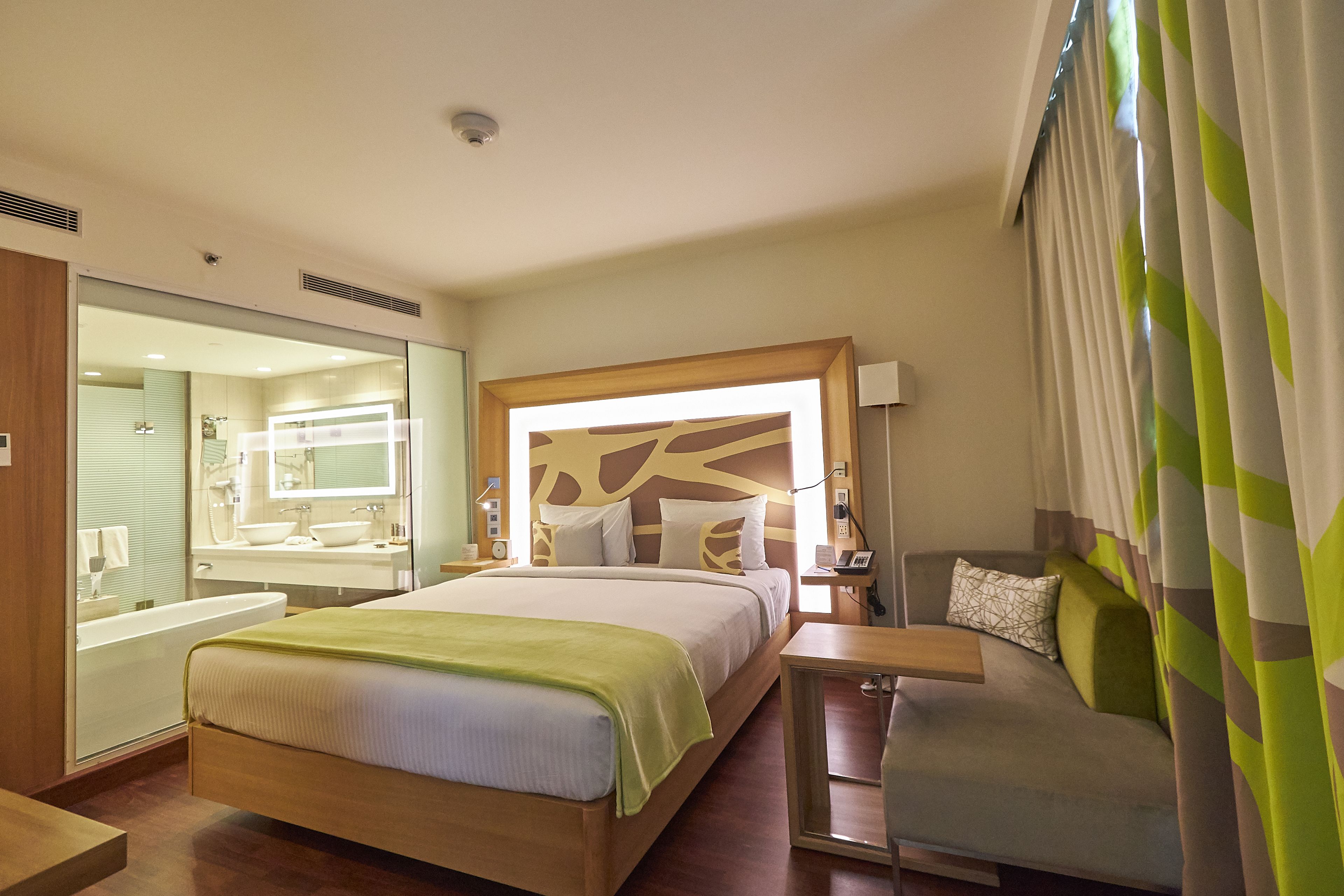 A hotel room with a bed, a sofa, a desk, a bathroom with a mirror, and a window with curtains.
