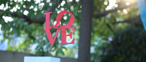 Una señal roja con la palabra 'LOVE' colgada al aire libre, sobre un fondo borroso de vegetación y luz solar.
