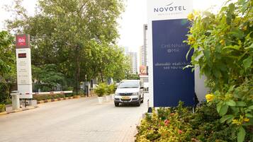 A hotel entrance with a sign for Novotel Hotels & Resorts, a white car parked in front, and a lush green garden with flowering plants.