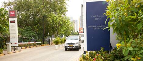 A hotel entrance with a sign for Novotel Hotels & Resorts, a white car parked in front, and a lush green garden with flowering plants.