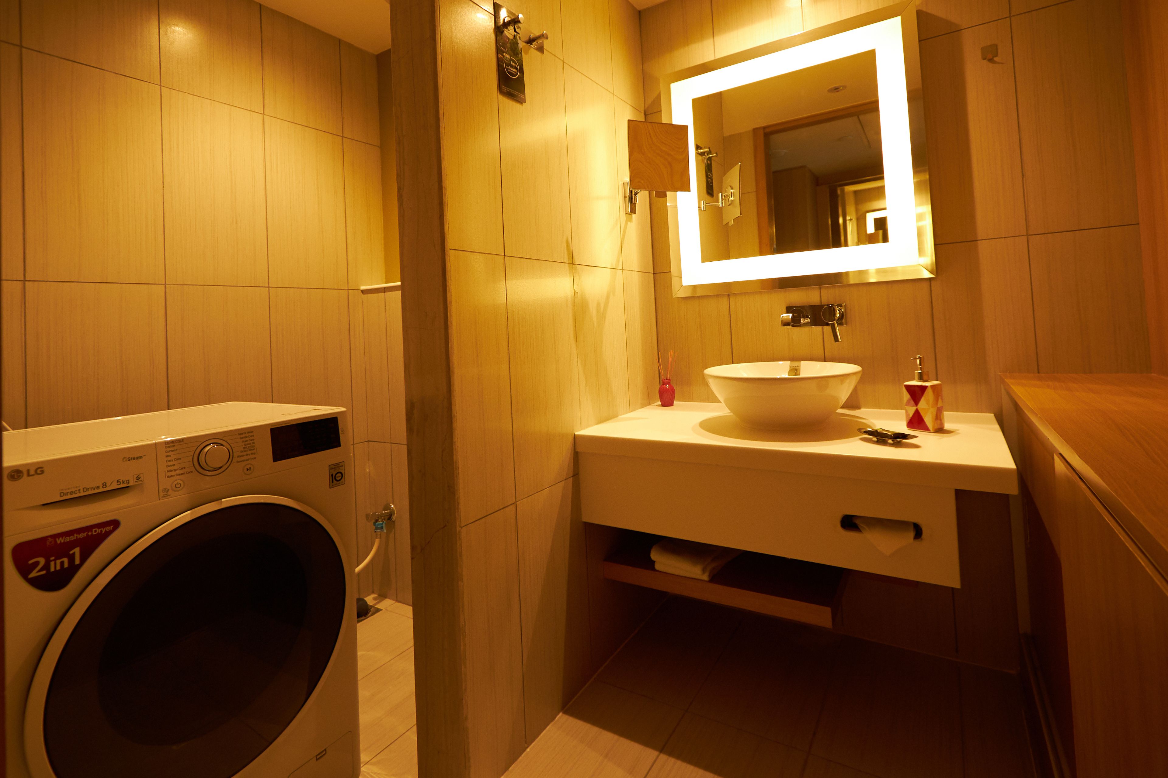 A compact hotel bathroom with a washing machine, a sink, and a mirror.