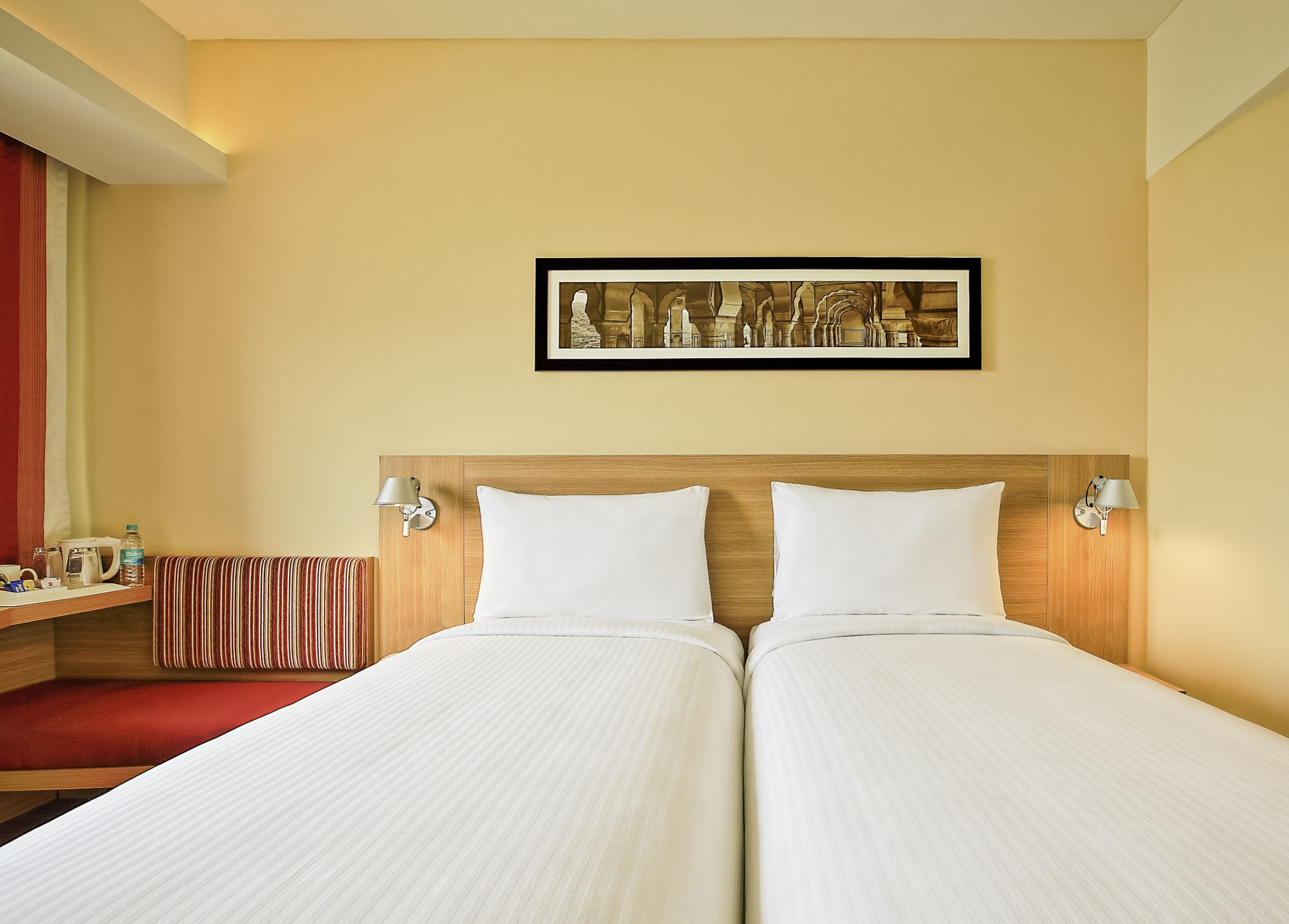 A hotel room with two beds, a wooden headboard, a framed artwork above the beds, bedside lamps, a red-striped bench, and a small table with a kettle and a bottle.