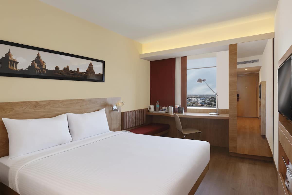 Ein Hotelzimmer mit einem großen Bett, einem Schreibtisch mit Stuhl, einem Fernseher und einem Fenster mit Blick auf die Stadt.