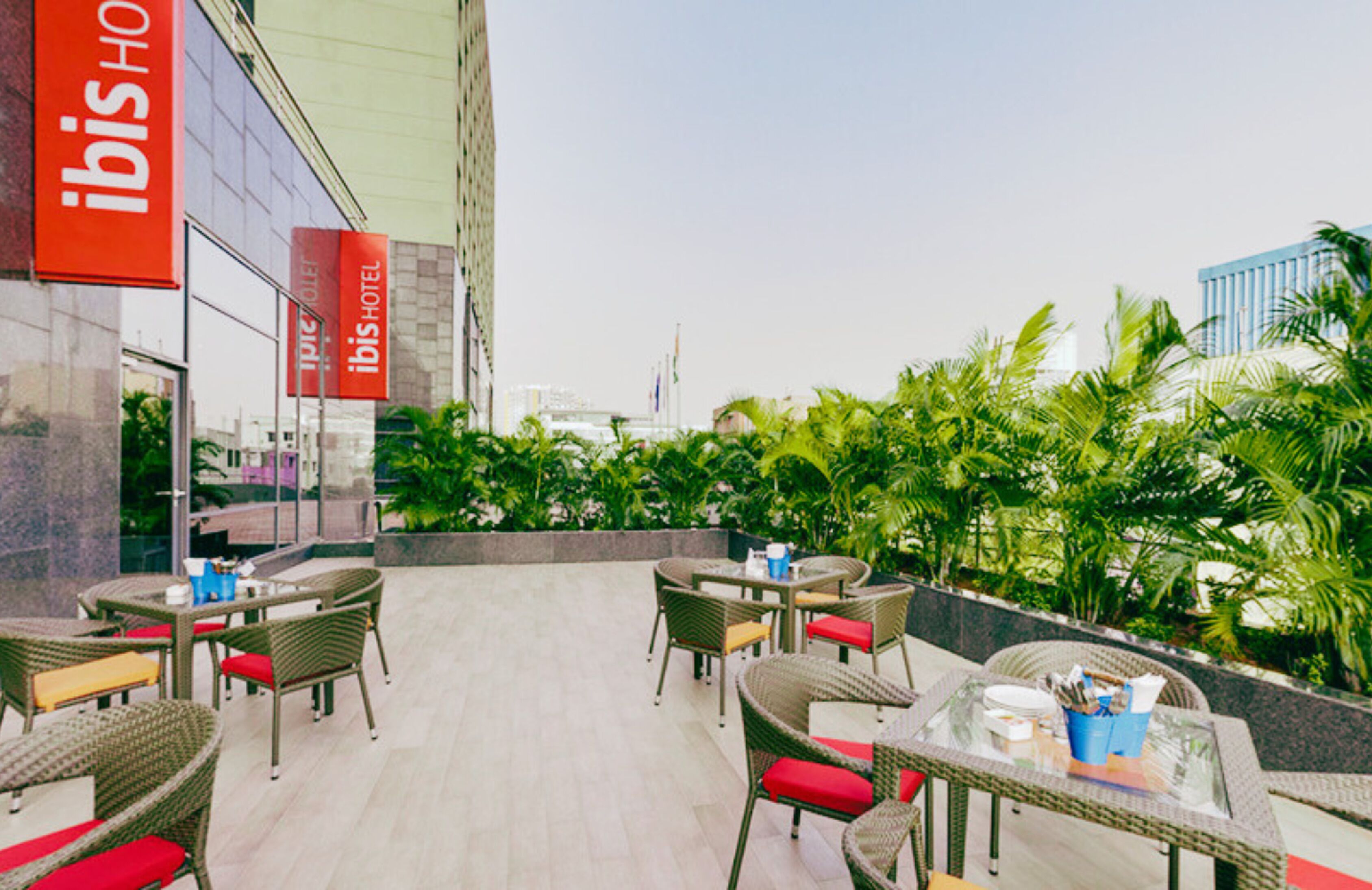 An outdoor seating area with wicker furniture and red cushions, surrounded by lush greenery.