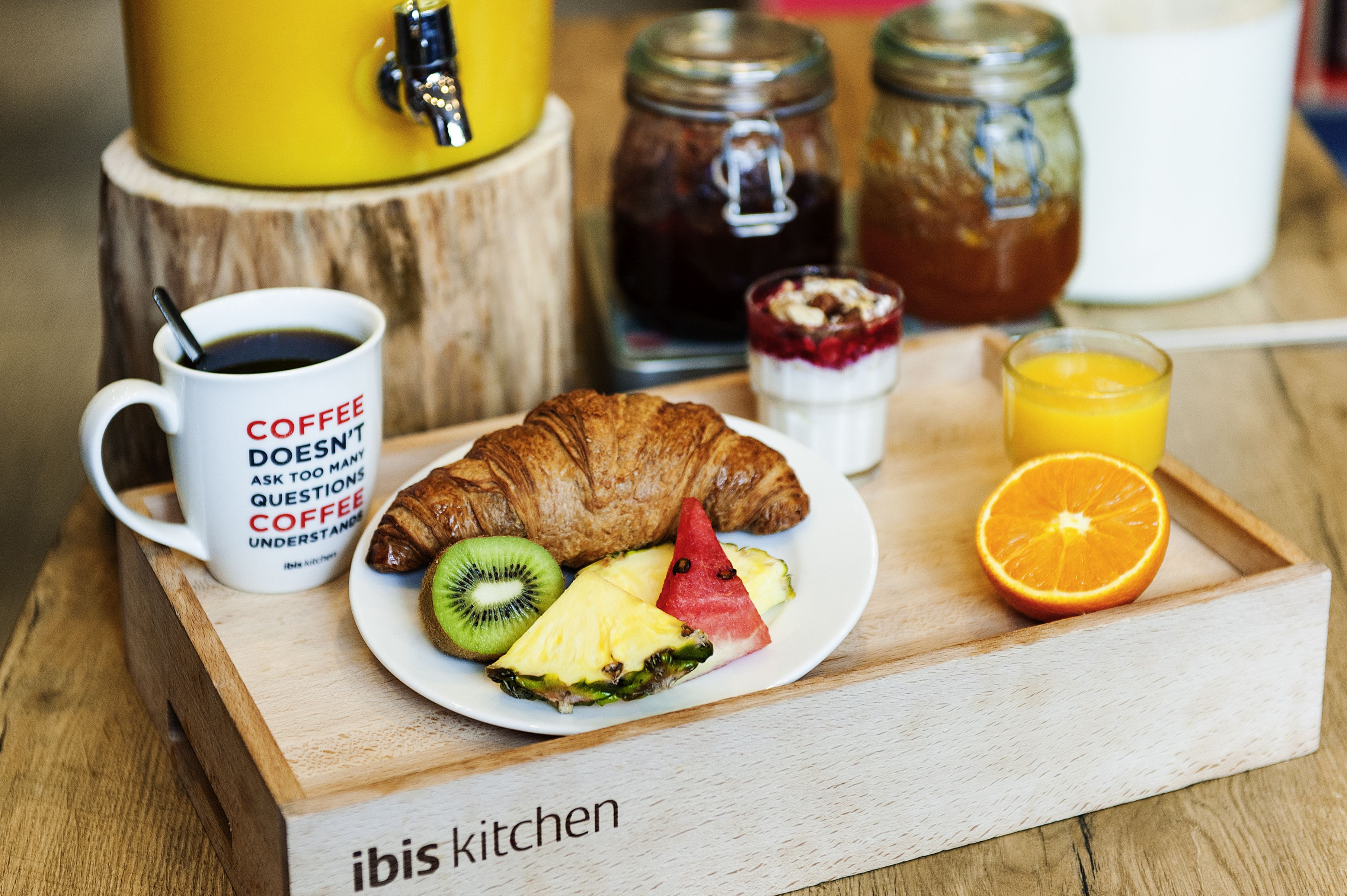 Bandeja de desayuno con croissant, kiwi, piña, sandía, jugo de naranja, café y un frasco de mermelada.