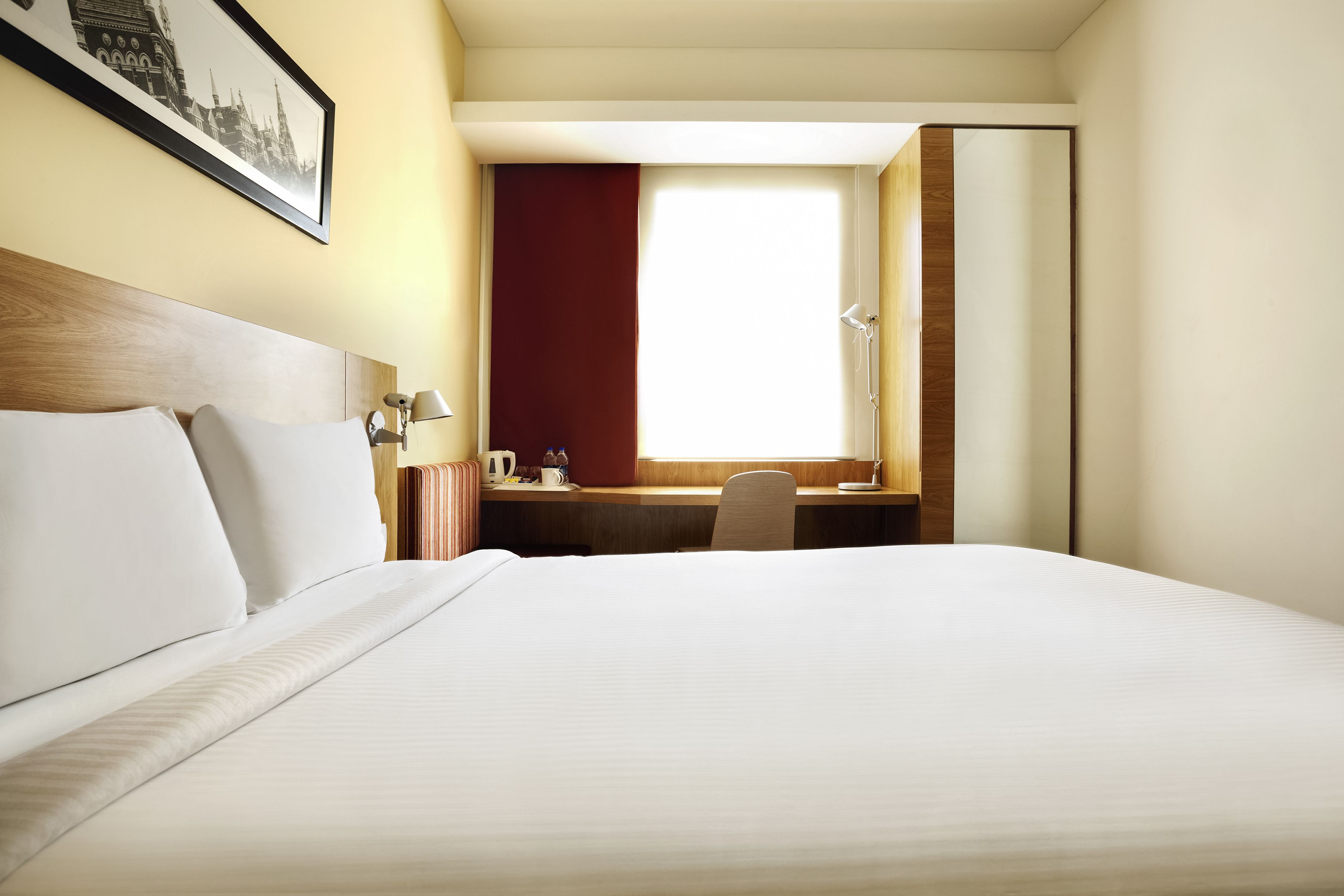 Ein Hotelzimmer mit einem großen Bett, einem Schreibtisch mit Stuhl, einem Fenster mit Vorhängen und einem gerahmten Bild an der Wand.