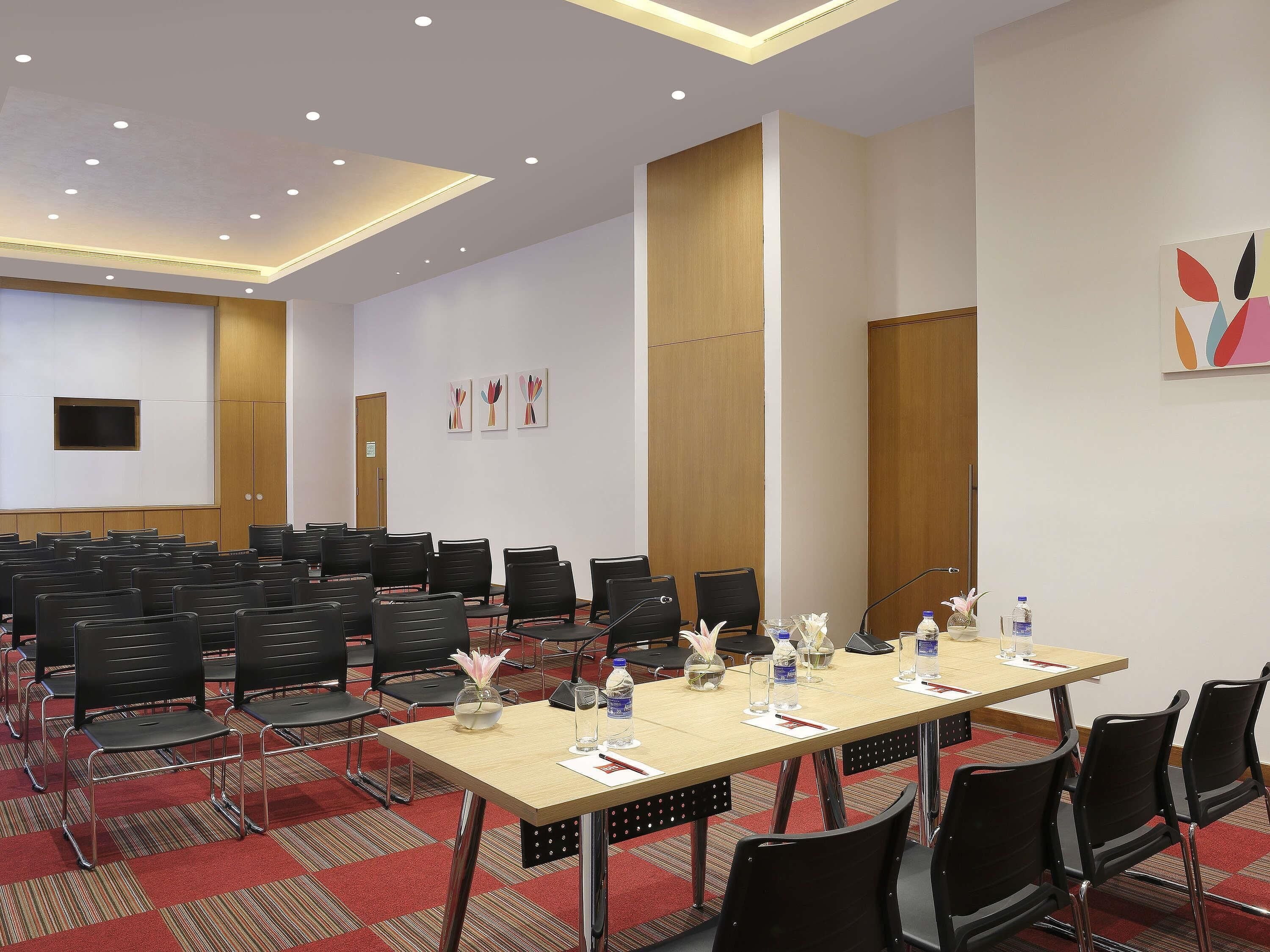A conference room with rows of chairs, a long table with microphones, and a television on the wall.