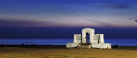 A coastal monument with a central archway and steps leading up to it, situated on a sandy beach with the sea in the background.