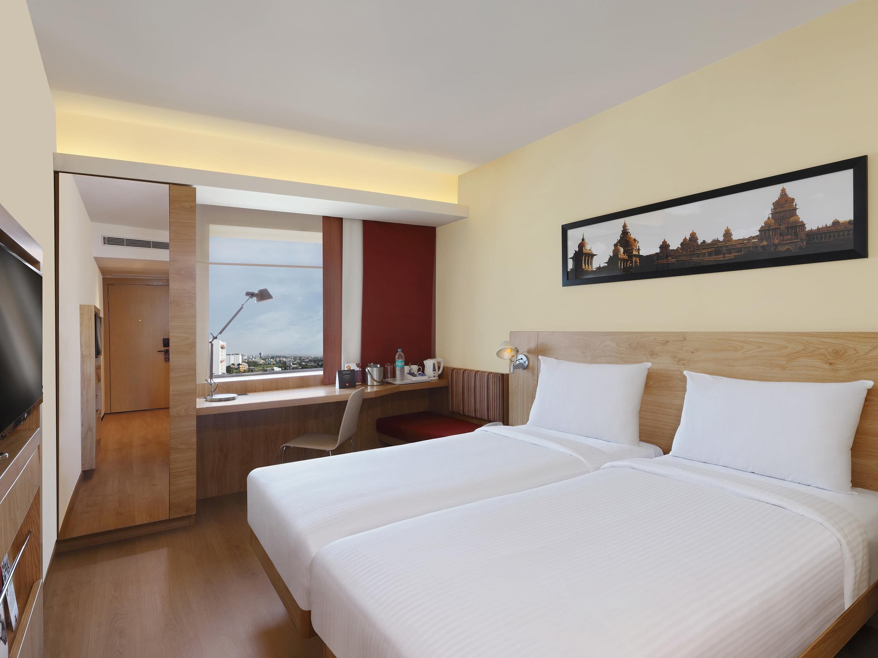 Ein Hotelzimmer mit zwei Betten, einem Schreibtisch, einem Fernseher und einem Fenster mit Blick auf die Stadt.