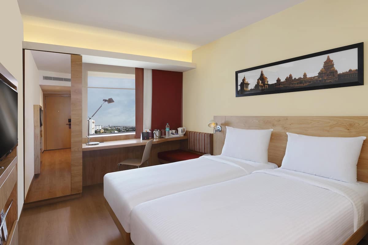Ein Hotelzimmer mit zwei Betten, einem Schreibtisch, einem Fernseher und einem Fenster mit Blick auf die Stadt.