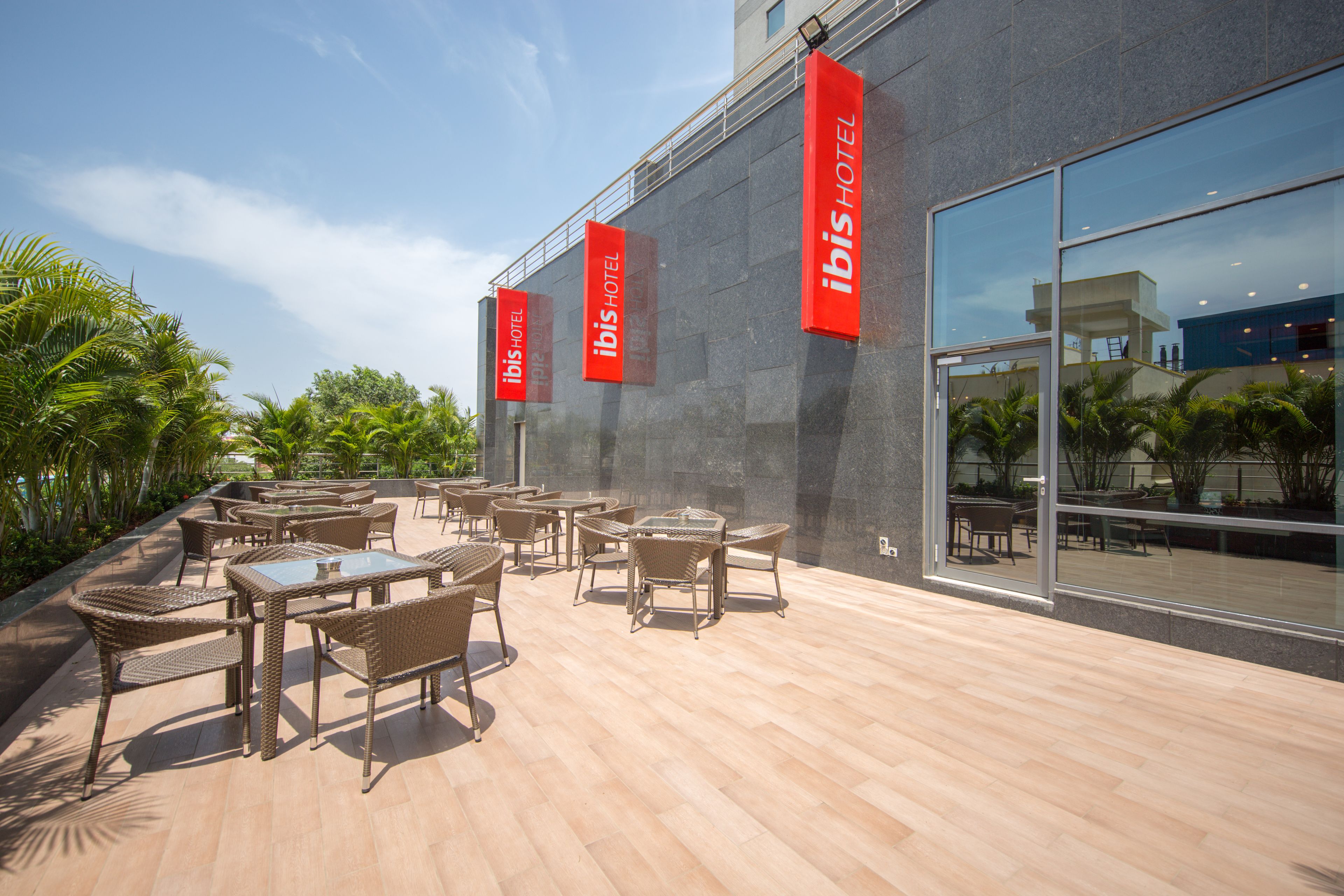 An outdoor seating area with wicker furniture and a glass wall reflecting the sky and palm trees.