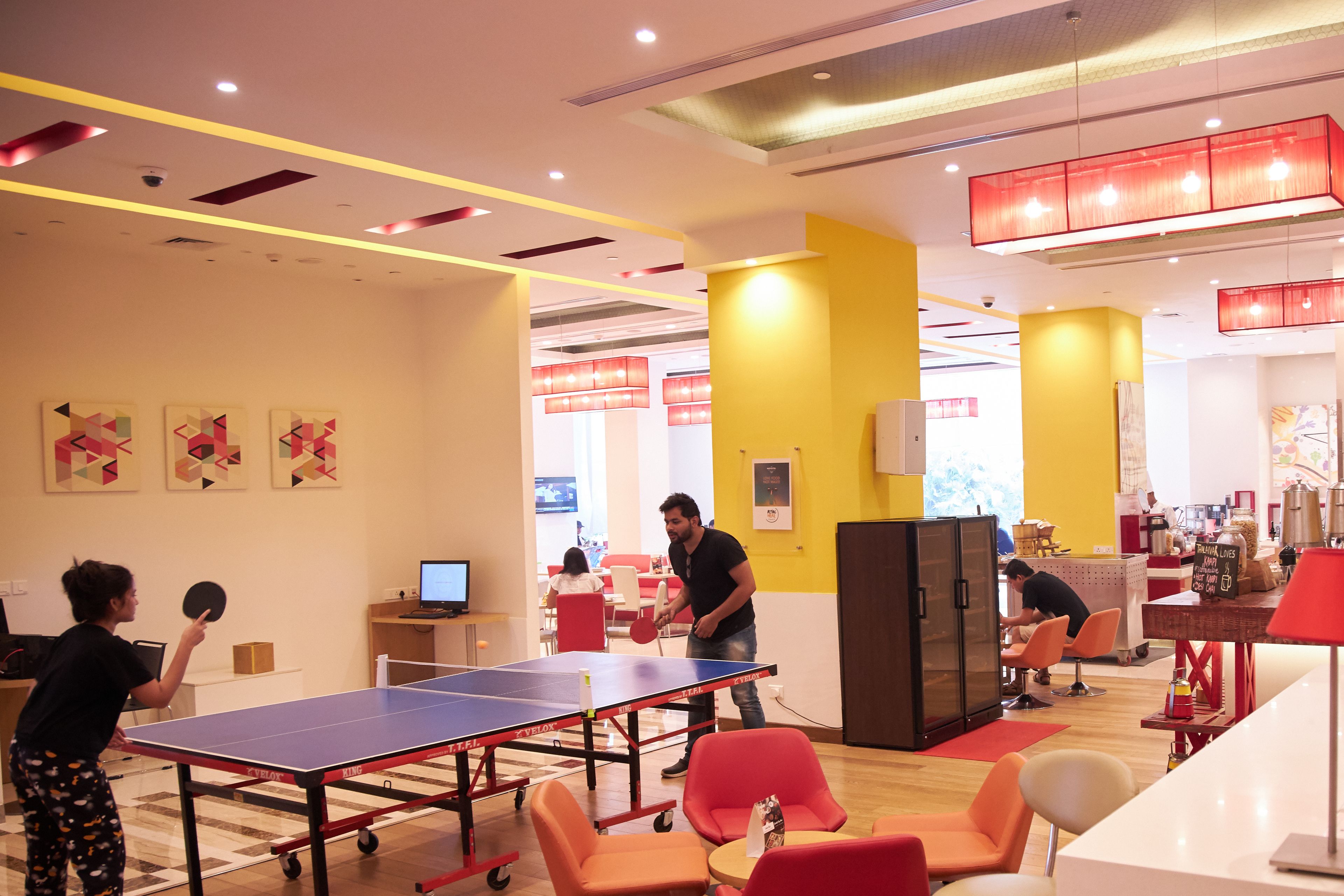 A ping pong table in a brightly lit room with yellow and white walls, a refrigerator, and several chairs.
