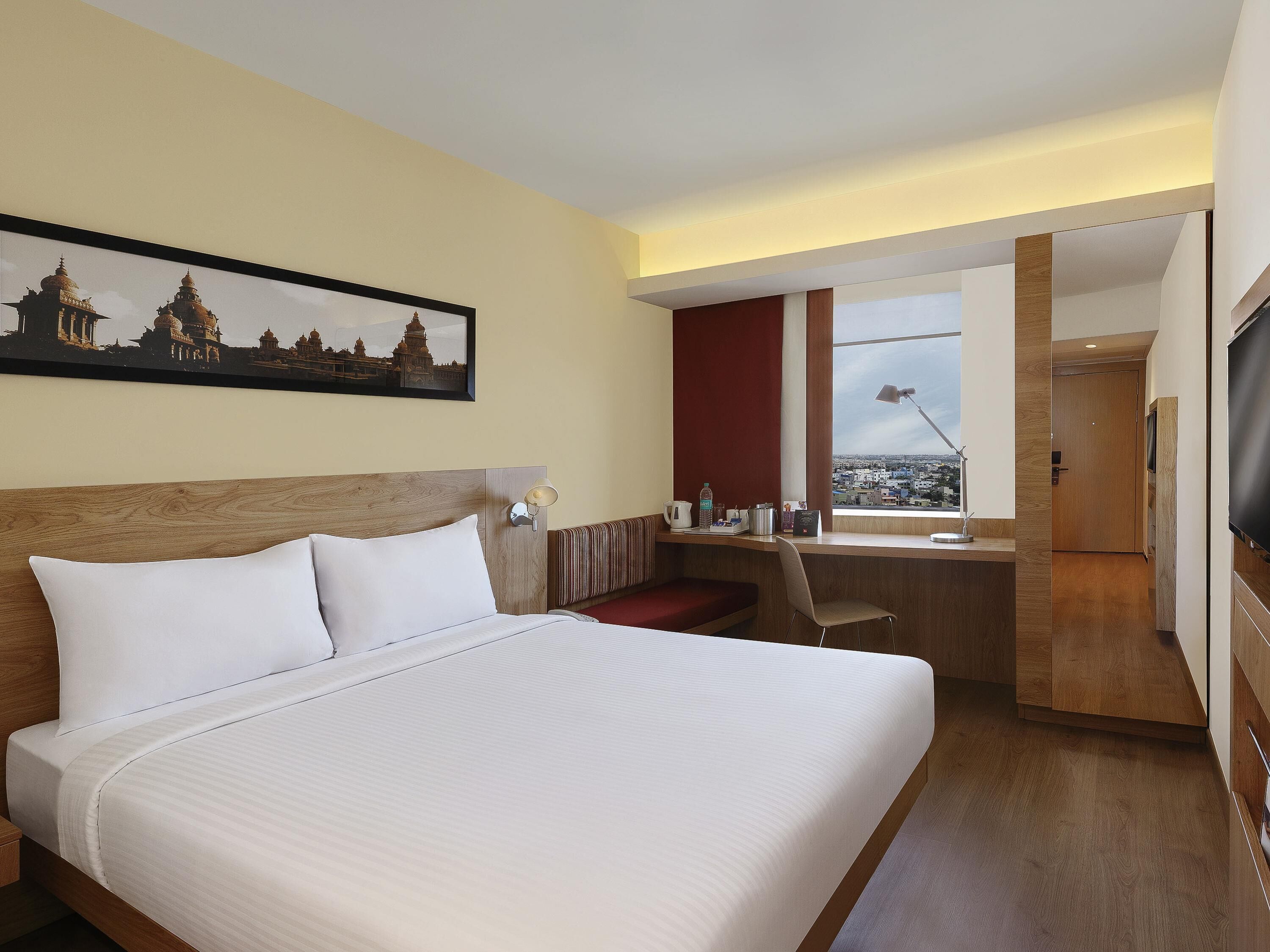 Ein Hotelzimmer mit einem großen Bett, einem Schreibtisch, einem Sessel, einem Fernseher und Blick auf die Stadt.