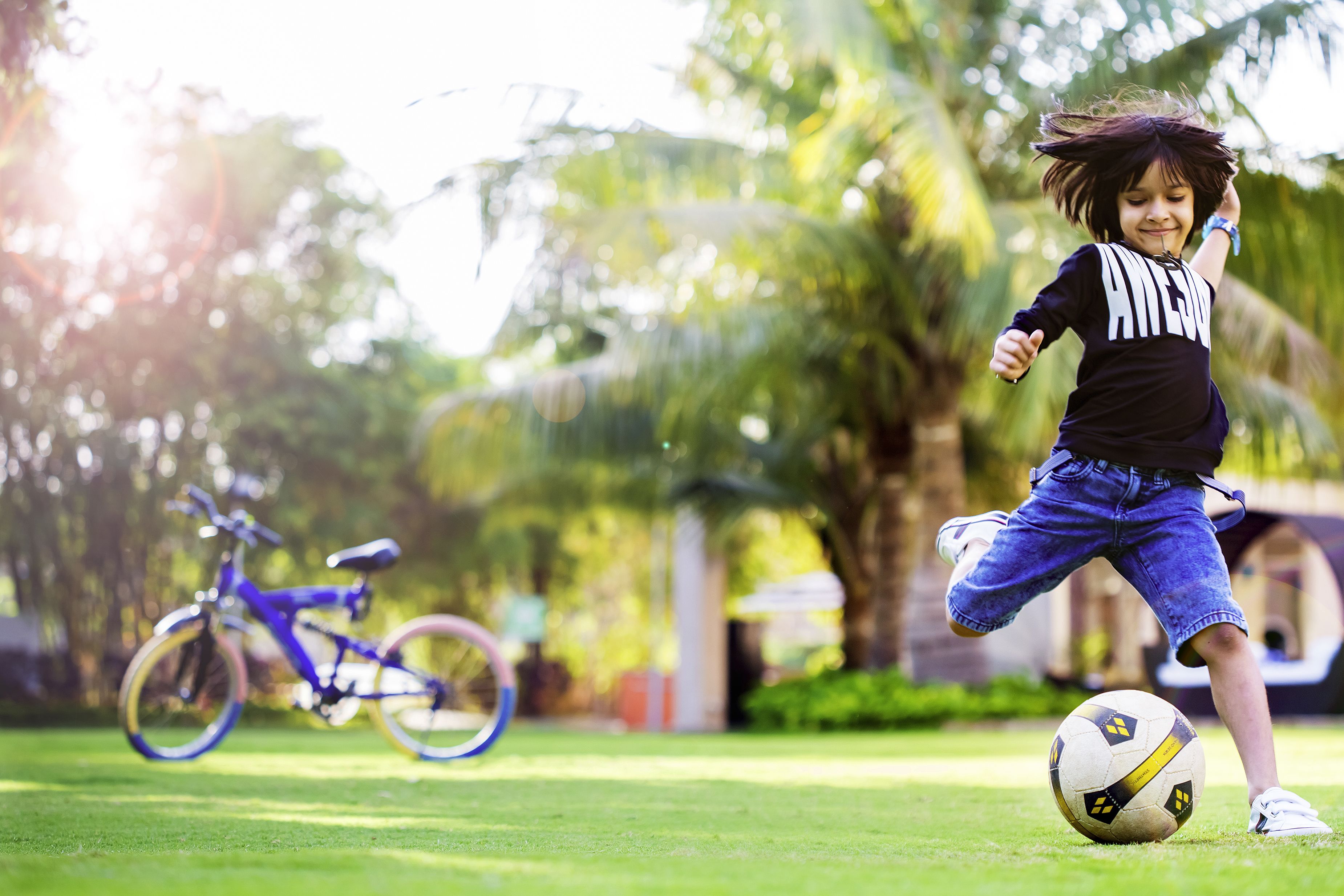 Niño jugando al fútbol en un campo cubierto de césped, con una bicicleta al fondo.