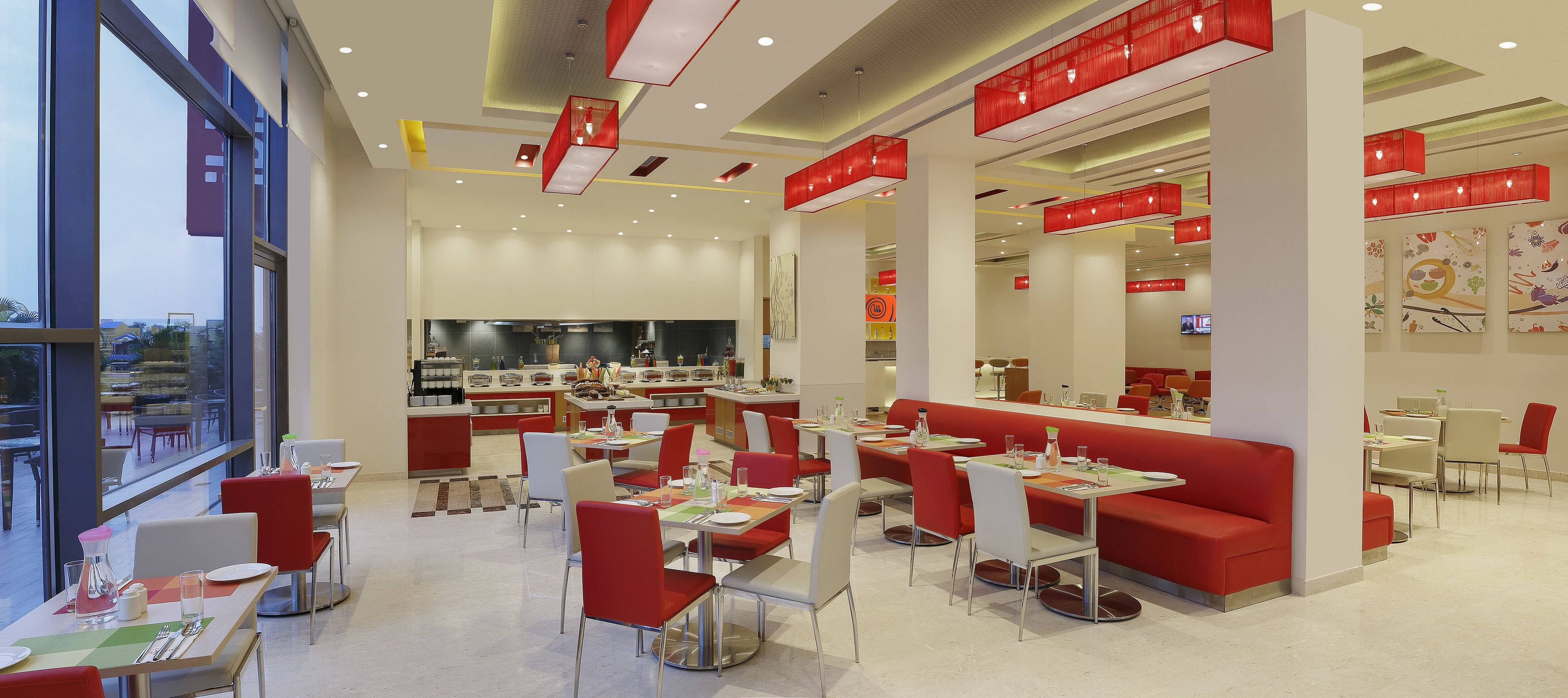 Un restaurant moderne à la décoration rouge et blanche, doté de plusieurs espaces de restauration et d’une cuisine ouverte visible au fond de la salle.