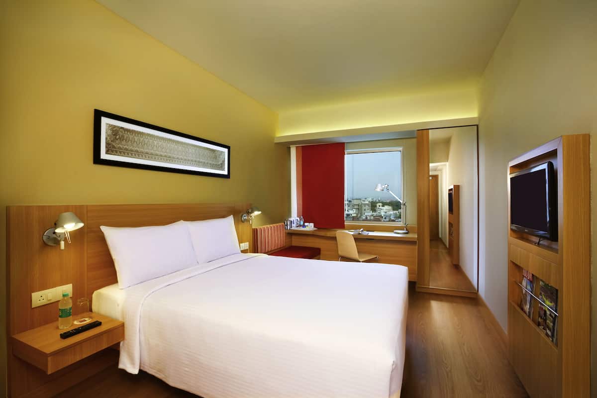Ein Hotelzimmer mit einem großen Bett, Nachttischlampen, einem Schreibtisch mit Stuhl, einem Fernseher und einem Fenster mit Blick auf die Stadt.