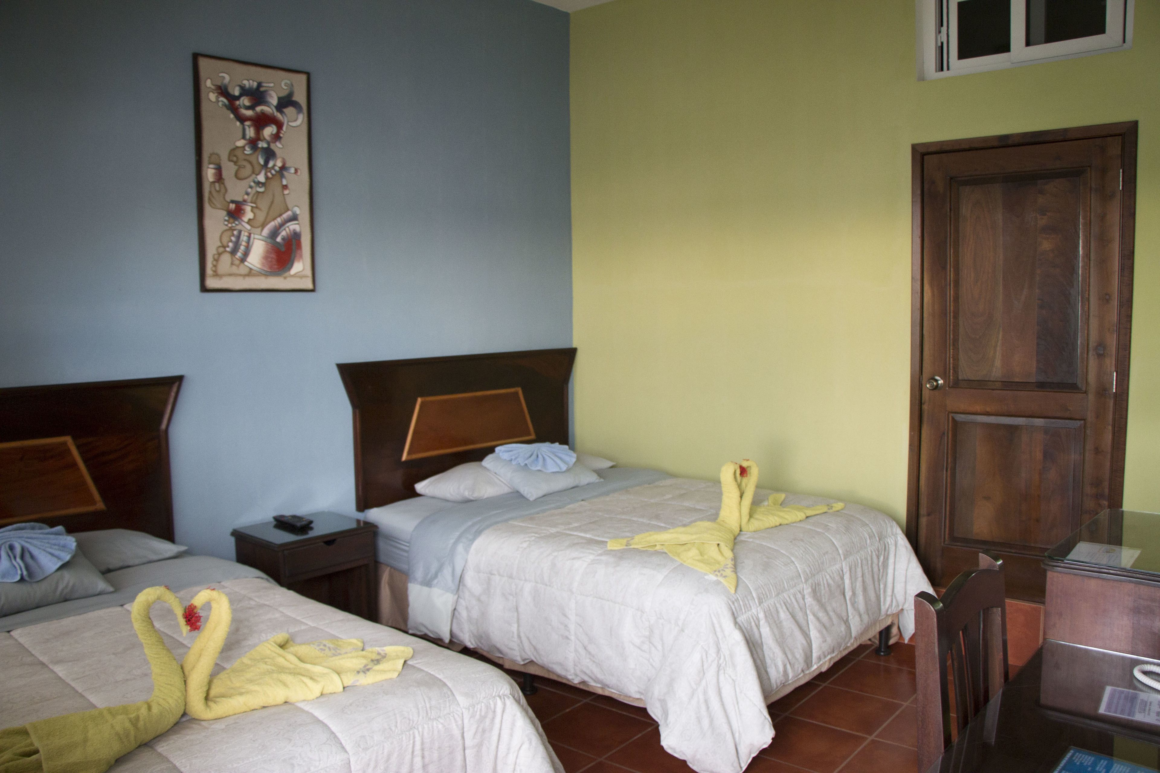 Quarto de hotel com duas camas, uma porta de madeira, uma mesa de cabeceira e um quadro na parede.