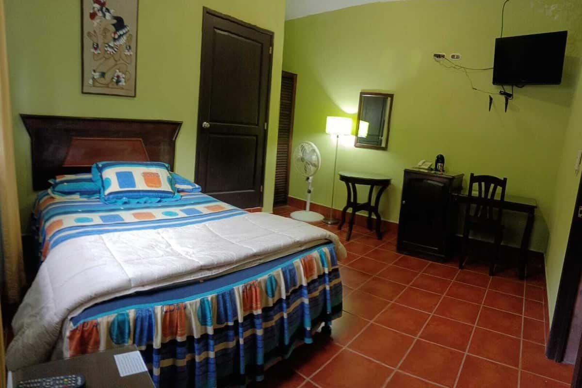 Quarto com cama, escrivaninha, cadeira, ventoinha e televisão.