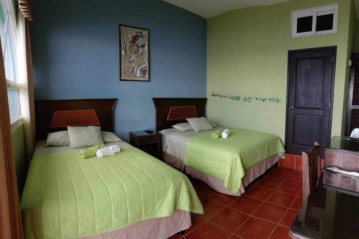 Quarto de hotel com duas camas, cada uma com colcha verde, cabeceiras de madeira e mesa de cabeceira com abajur.