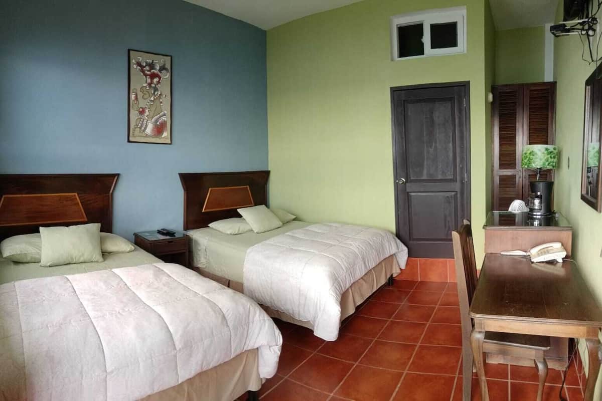 Quarto com duas camas, uma mesa de madeira, uma cadeira, uma porta e uma janela pequena.