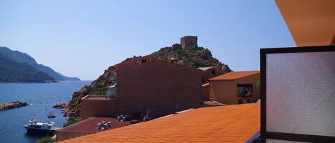 A coastal building with a red-tiled roof overlooking a body of water and a hill with a tower.