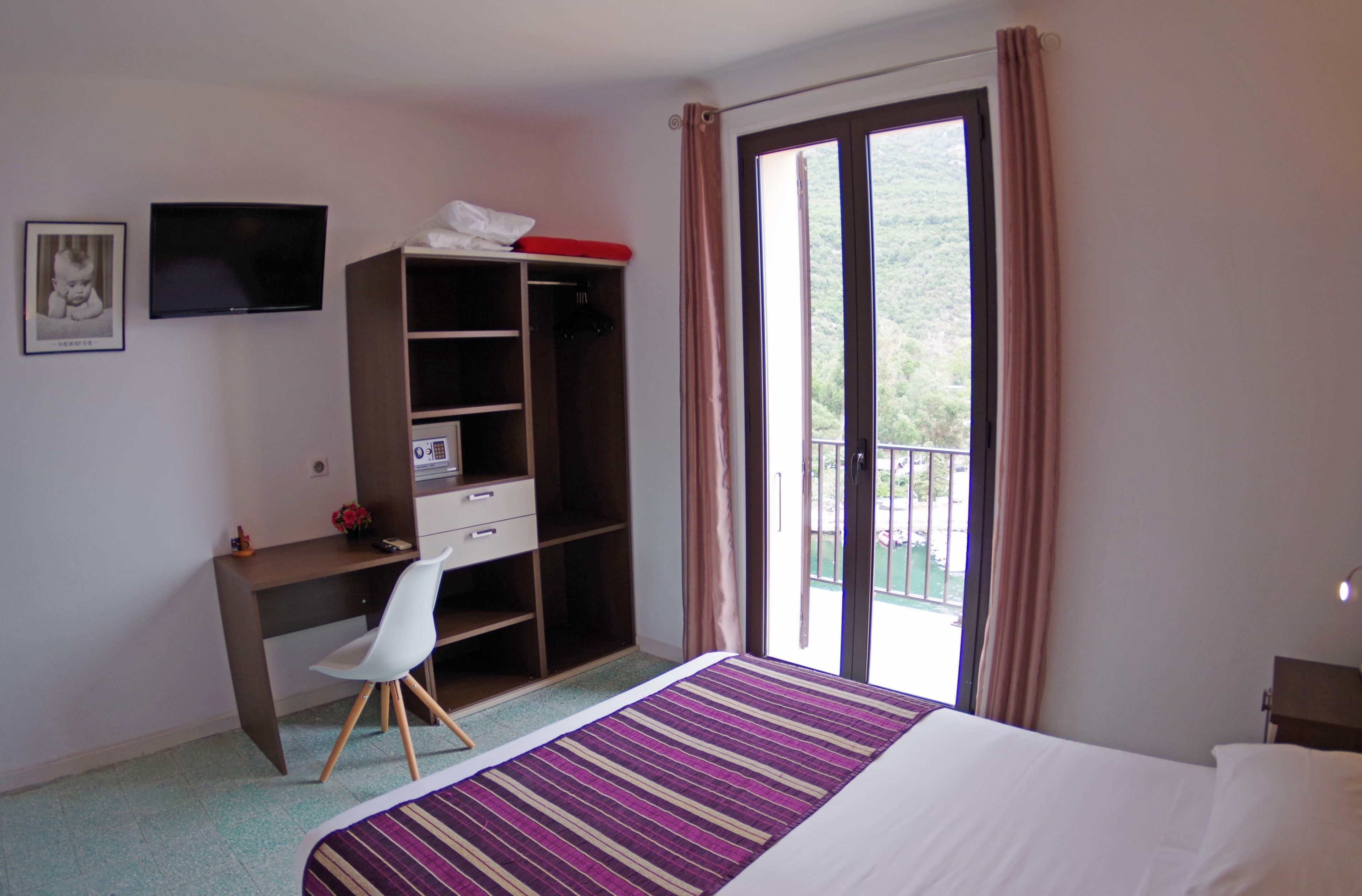 A hotel room with a bed, a desk, a chair, a wardrobe, a television, and a balcony with curtains.