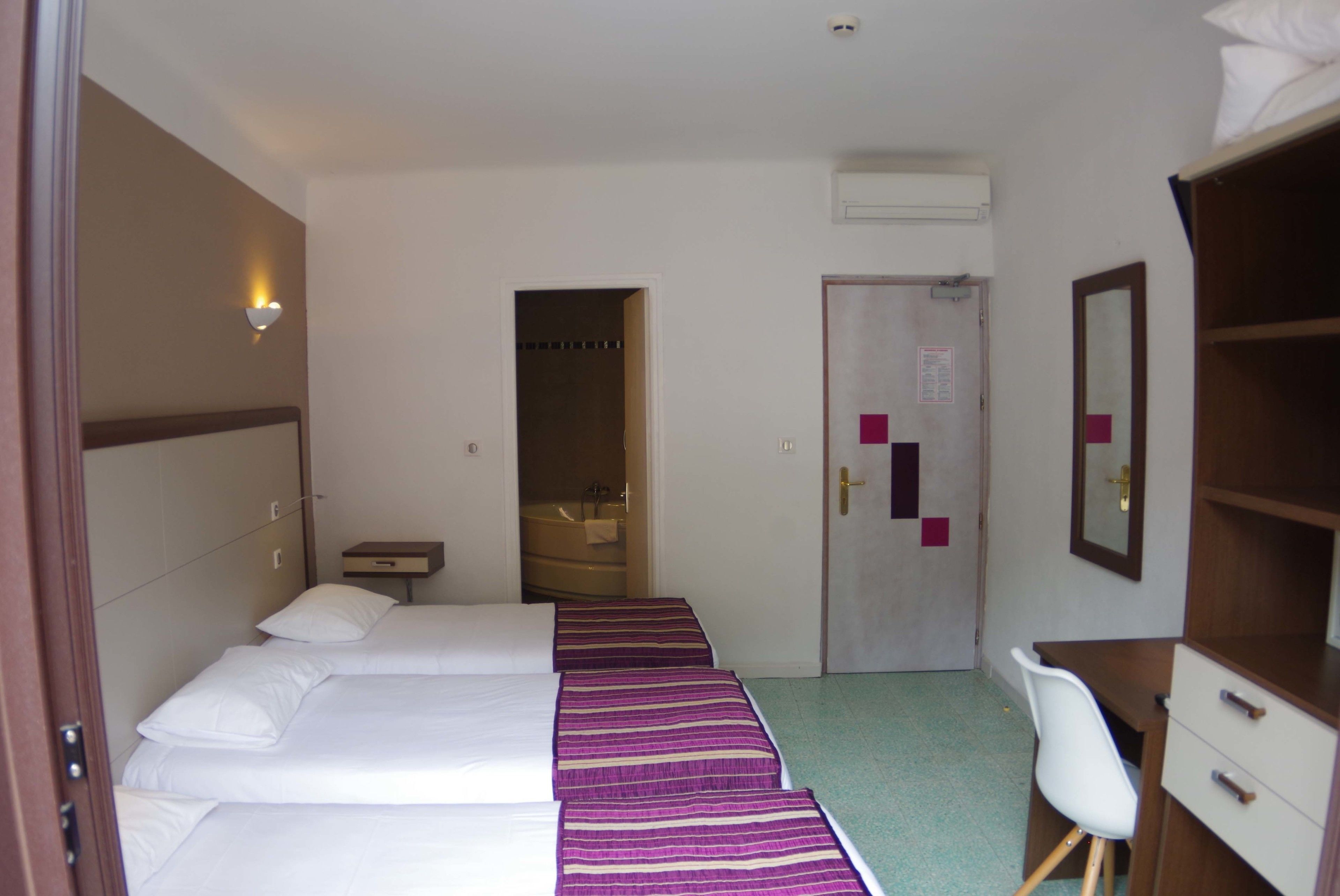 A hotel room with three beds, a desk, a chair, and a bathroom visible through an open door.