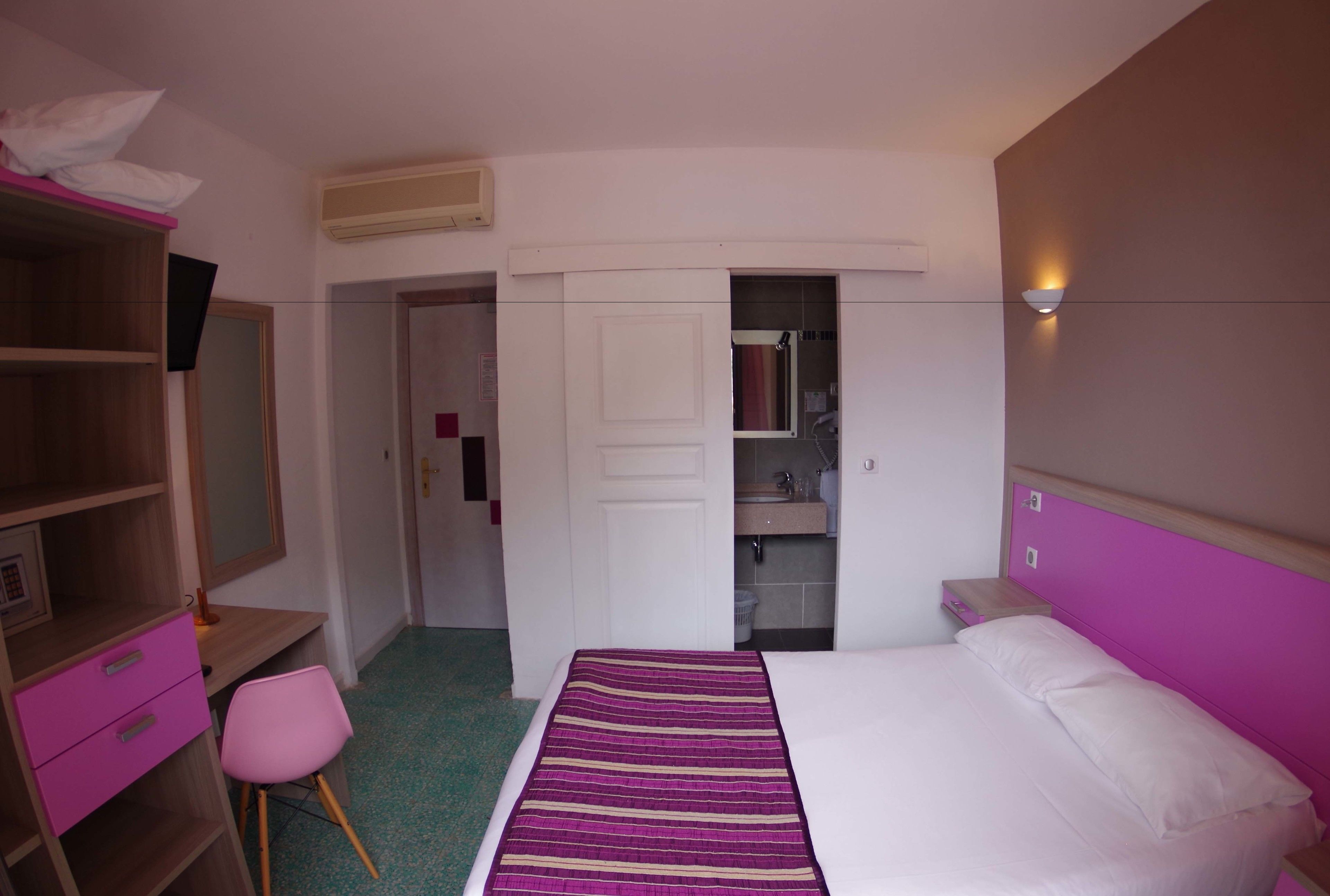 A bedroom with a bed, a desk, a chair, a wardrobe, and a bathroom visible through an open door.