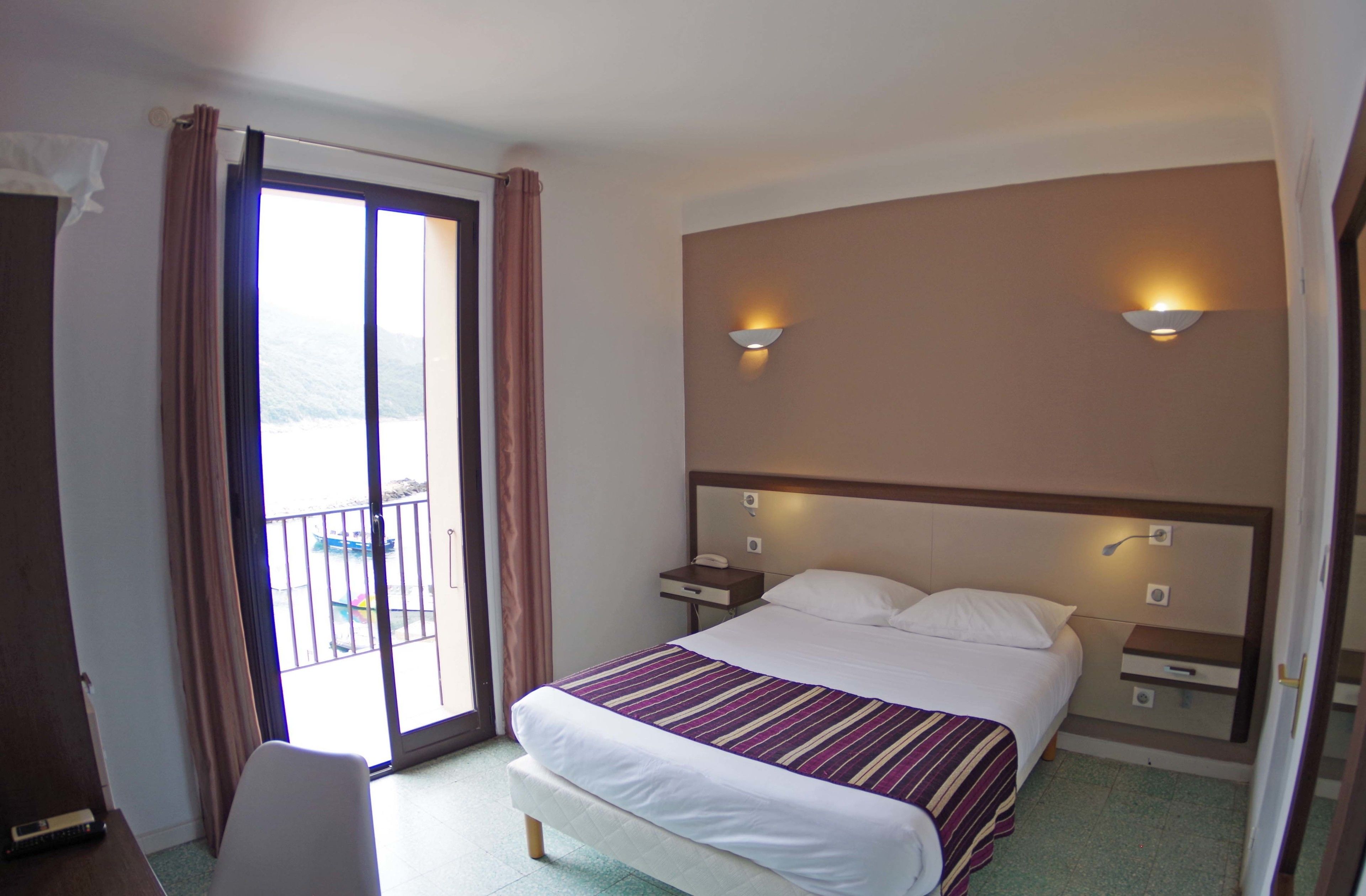 A hotel room with a bed, a nightstand, a chair, a balcony with a view, and two wall-mounted lights.