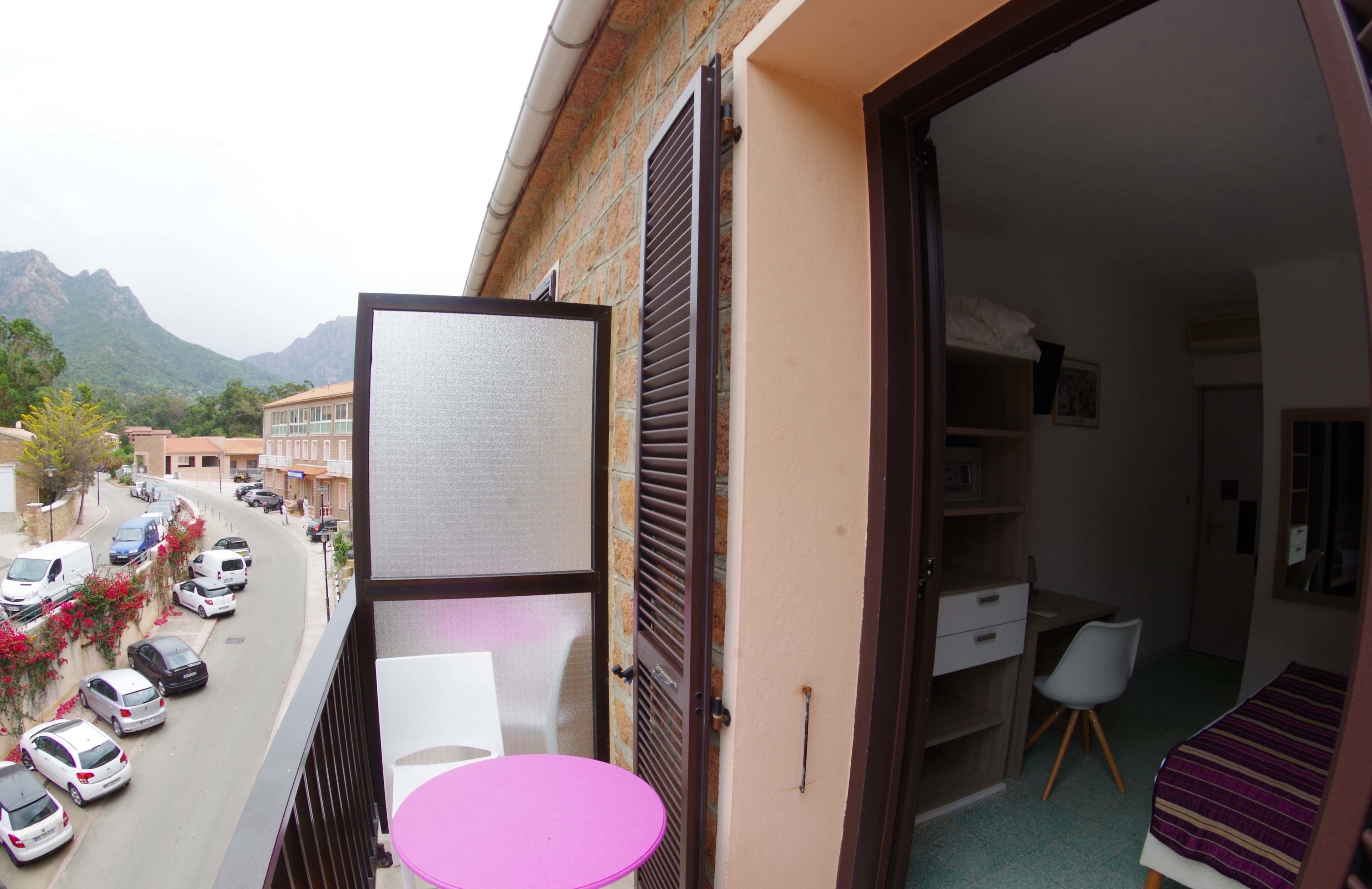 Un balcón con vista a una calle y montañas, una mesa rosa y un dormitorio con cama y escritorio.