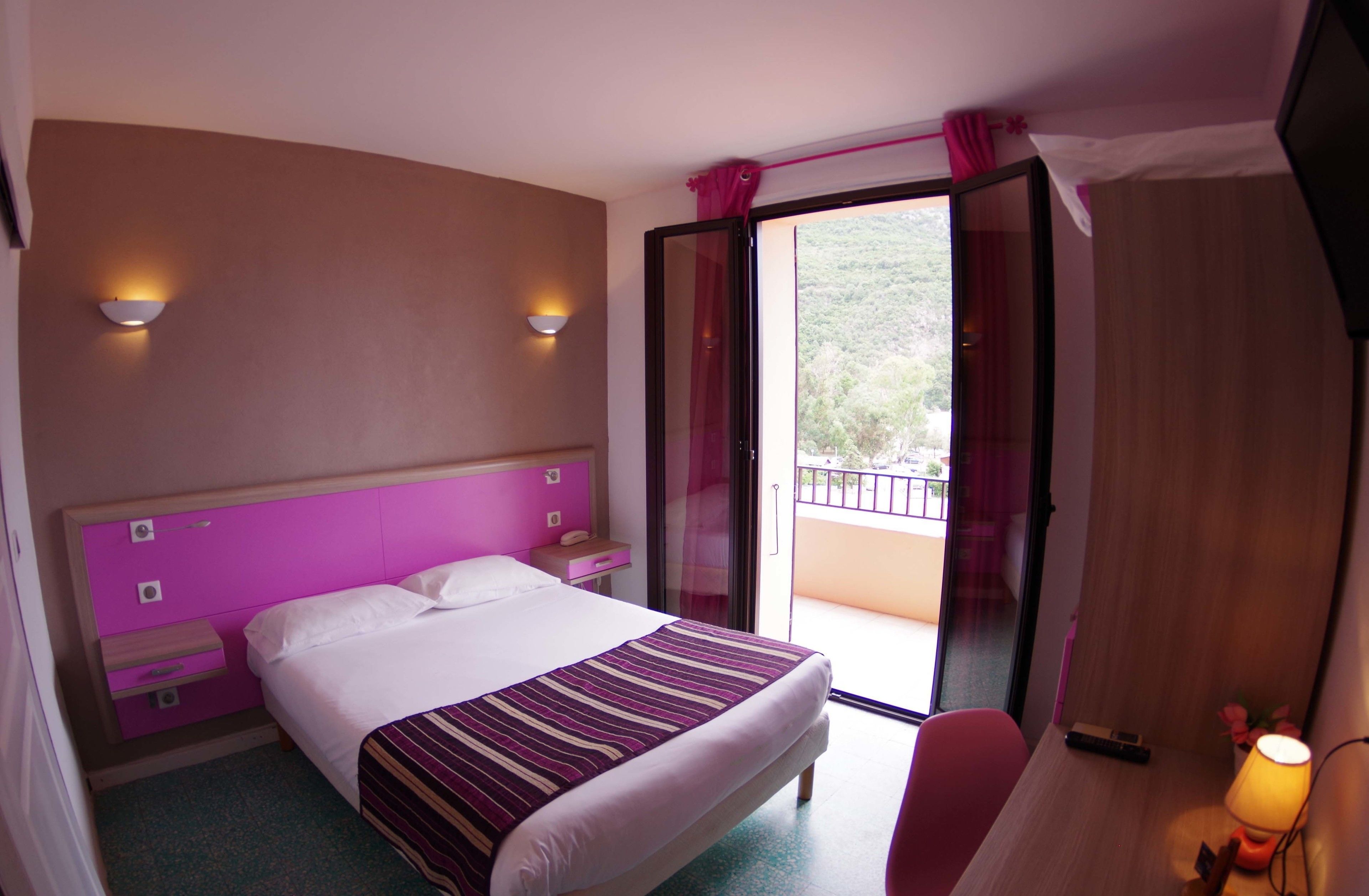A hotel room with a bed, a desk, a chair, a nightstand, a lamp, and a sliding glass door leading to a balcony.