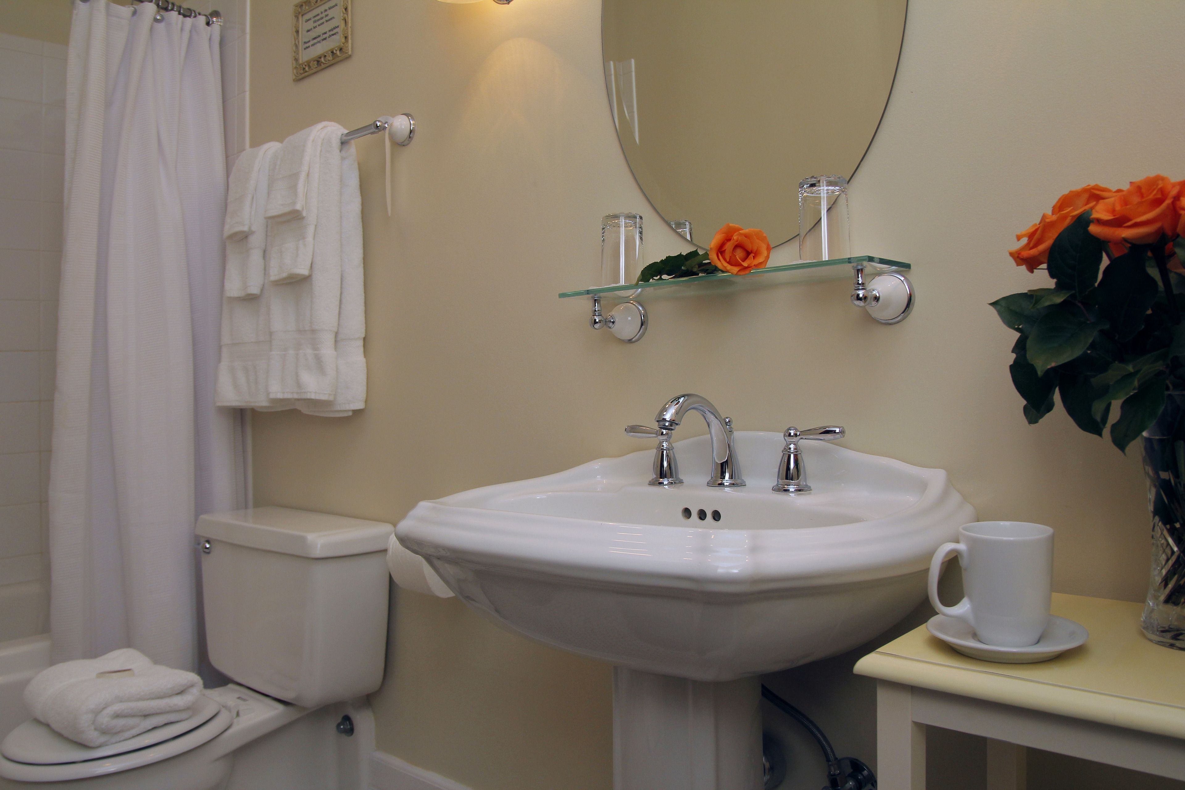 A bathroom with a white sink, a toilet, a glass shelf with a rose, and a white cup on a small table.