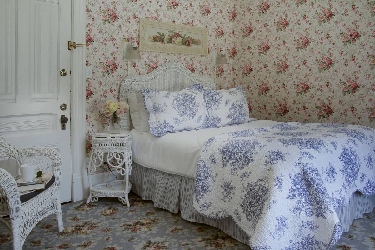 A bedroom with a floral wallpaper, a white bed with blue floral bedding, a wicker chair, and a small bedside table with a lamp.