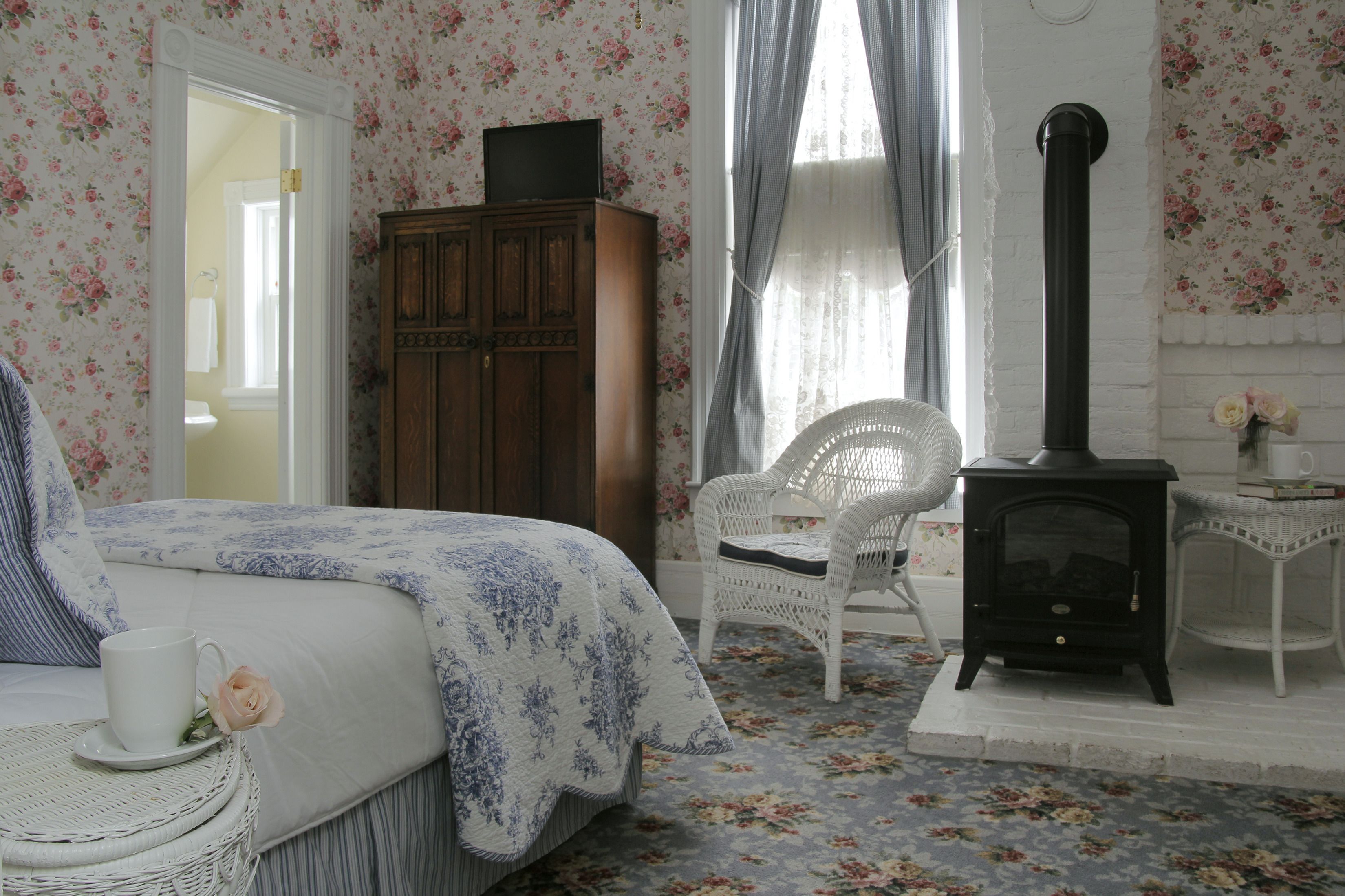 A bedroom with floral wallpaper, a bed with a blue and white bedspread, a wicker chair, a white cabinet, and a black stove.