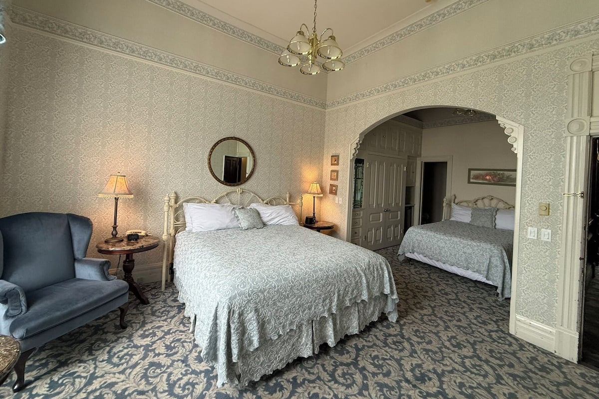 A hotel room with two beds, a chandelier, a mirror, and a blue armchair.