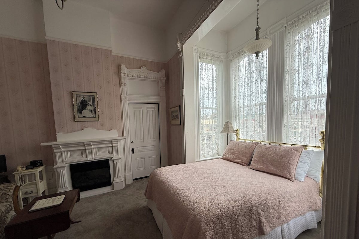 A bedroom with a bed, a fireplace, a desk, and a window with curtains.