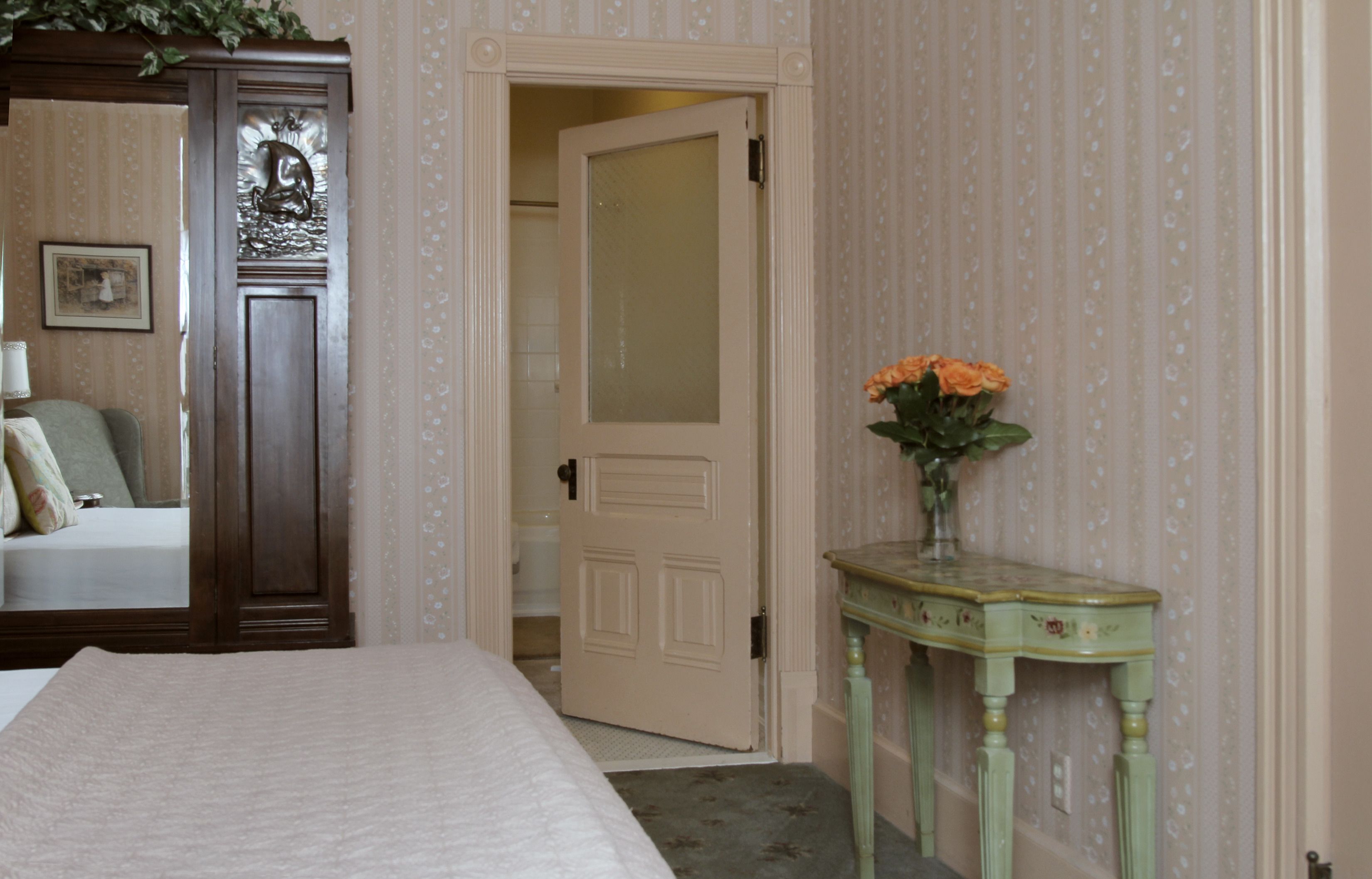 Un dormitorio con cama, cómoda, espejo, puerta y una mesita con un florero.